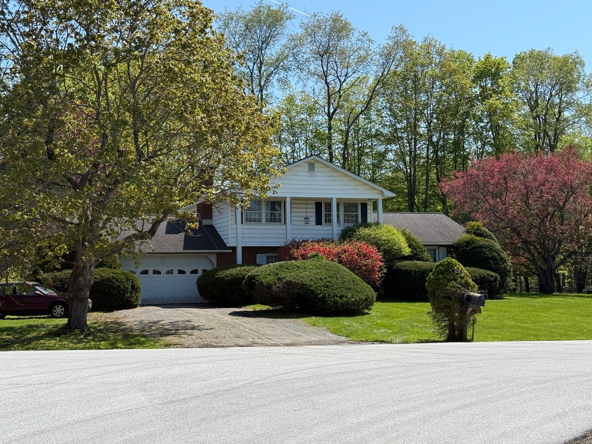 White two-story house with attached garage, surrounded by green grass, bushes, and trees.