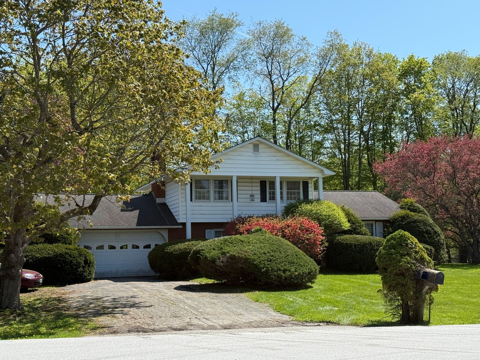 Two-story white house with attached garage, surrounded by green shrubs, trees, and a driveway.
