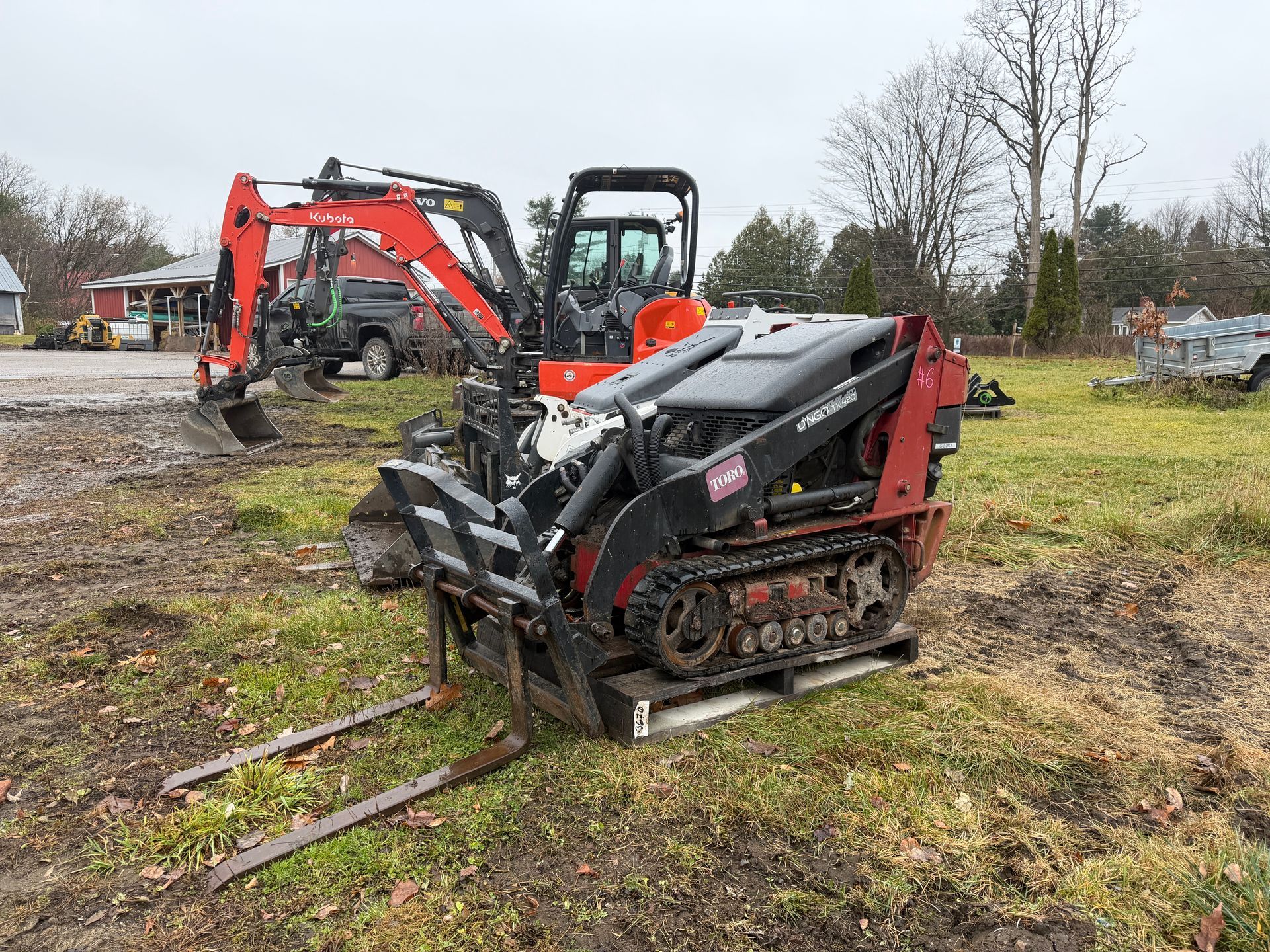 A track skid steer with forks, in front of a red excavator on a grassy field.