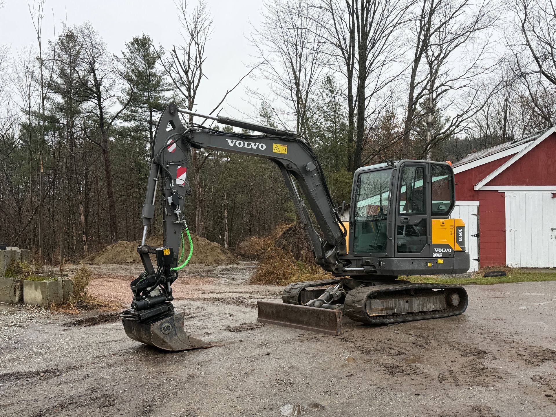 Volvo excavator on a muddy surface, trees in the background, a red building to the right.