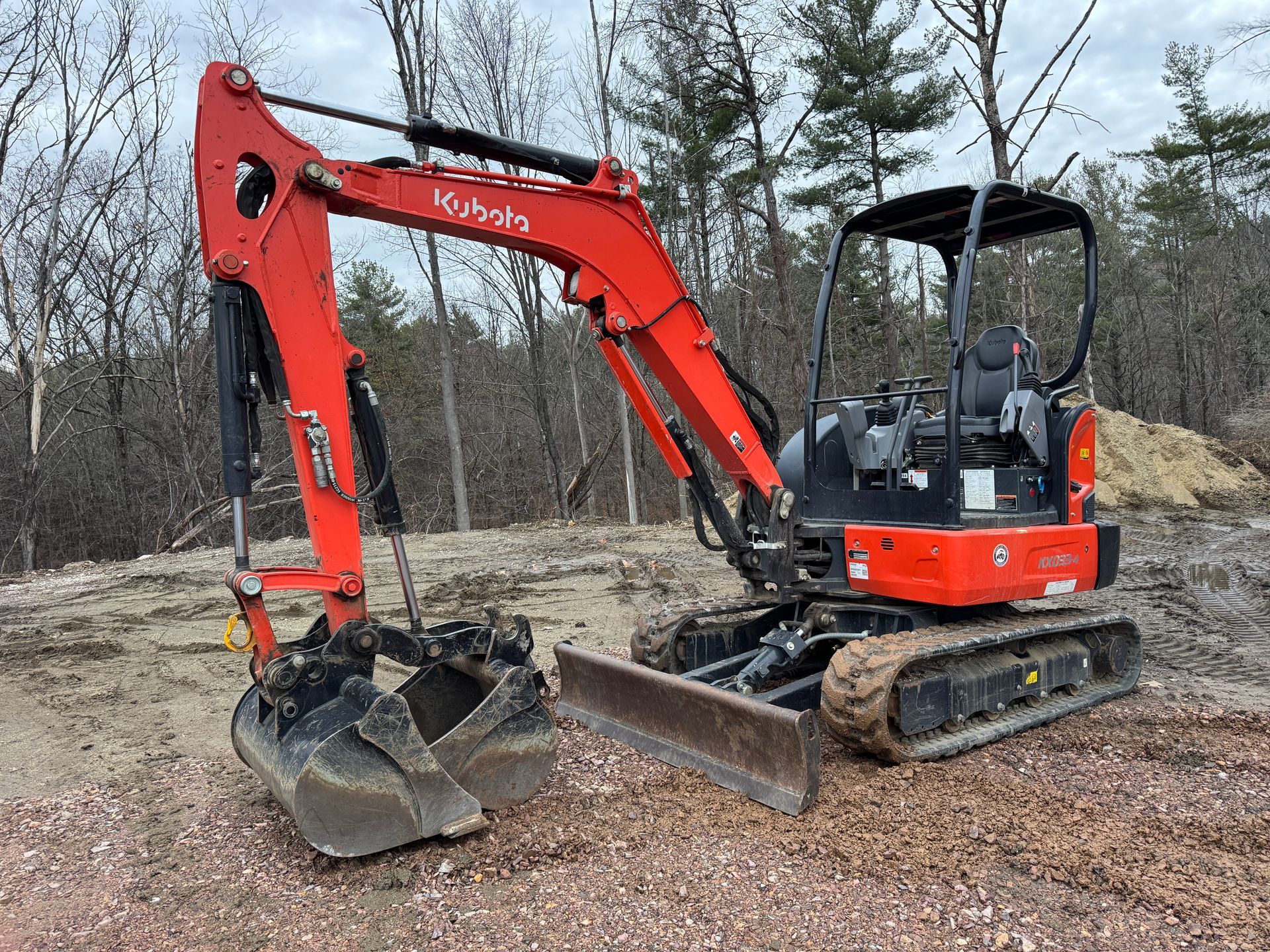 Orange Kubota excavator with a bucket and blade, parked on gravel in front of trees.