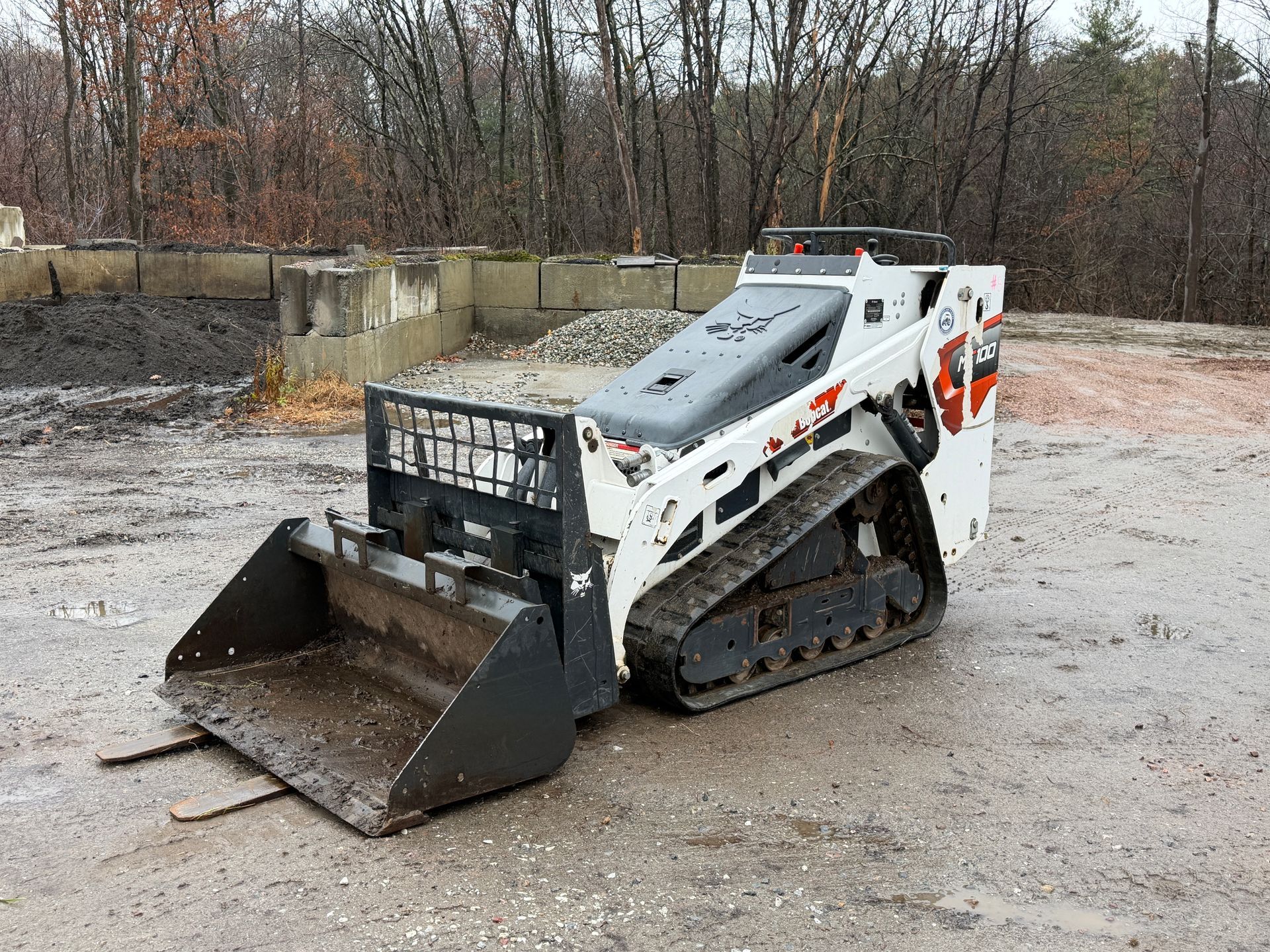 White and orange Bobcat compact track loader with bucket and forks on wet ground.