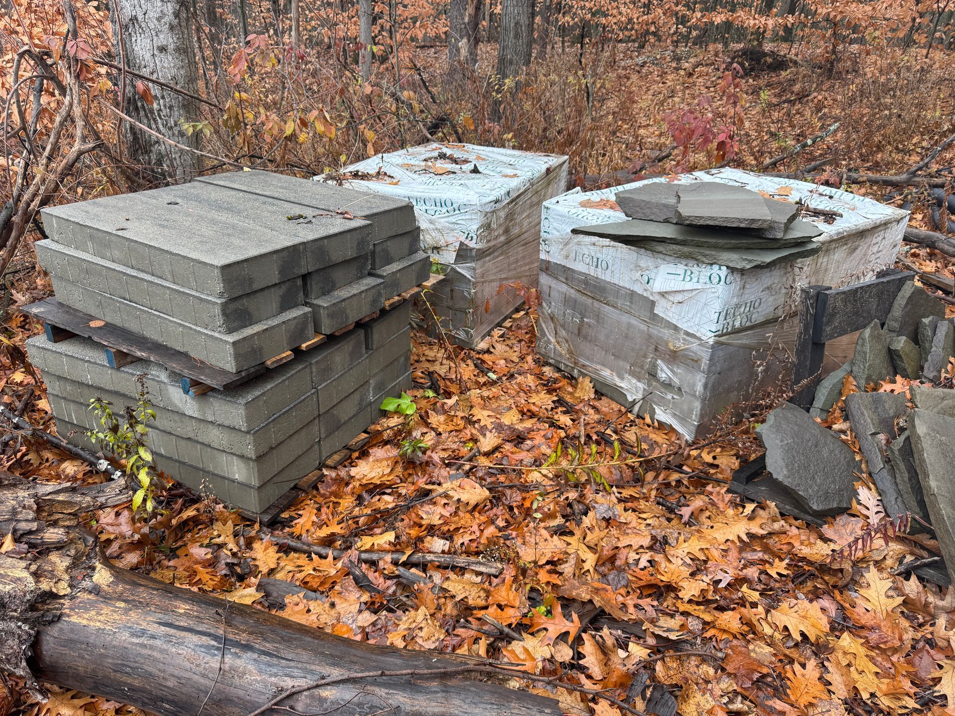 Three stacks of gray and white concrete blocks on fallen leaves in a wooded area.