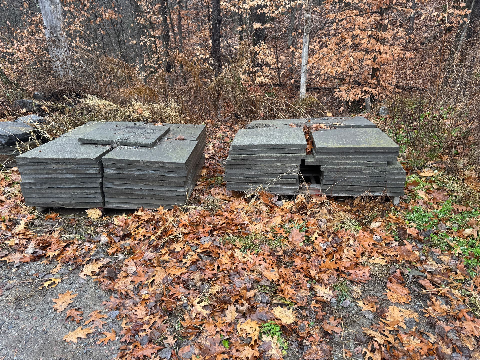 Two stacks of gray square paving stones in a wooded area with fallen autumn leaves.