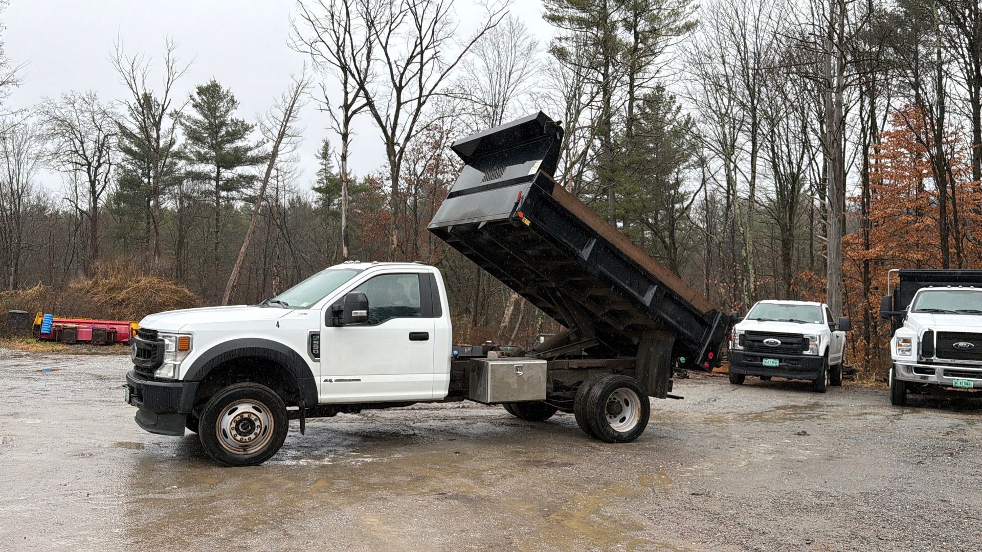 White dump truck with raised bed, parked on gravel, with two other trucks in the background.