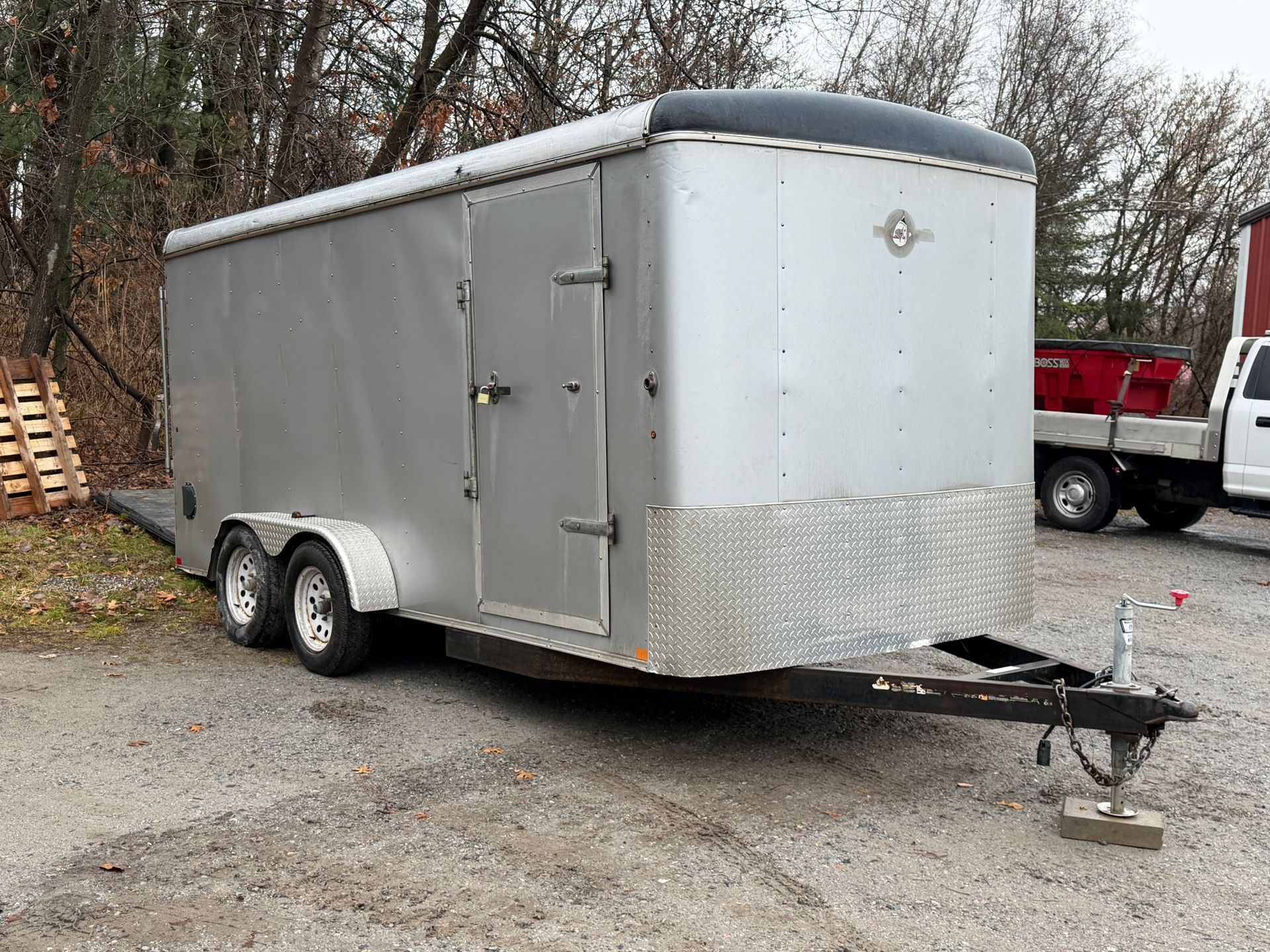 Silver enclosed trailer with black top and diamond plate lower front, parked on gravel.