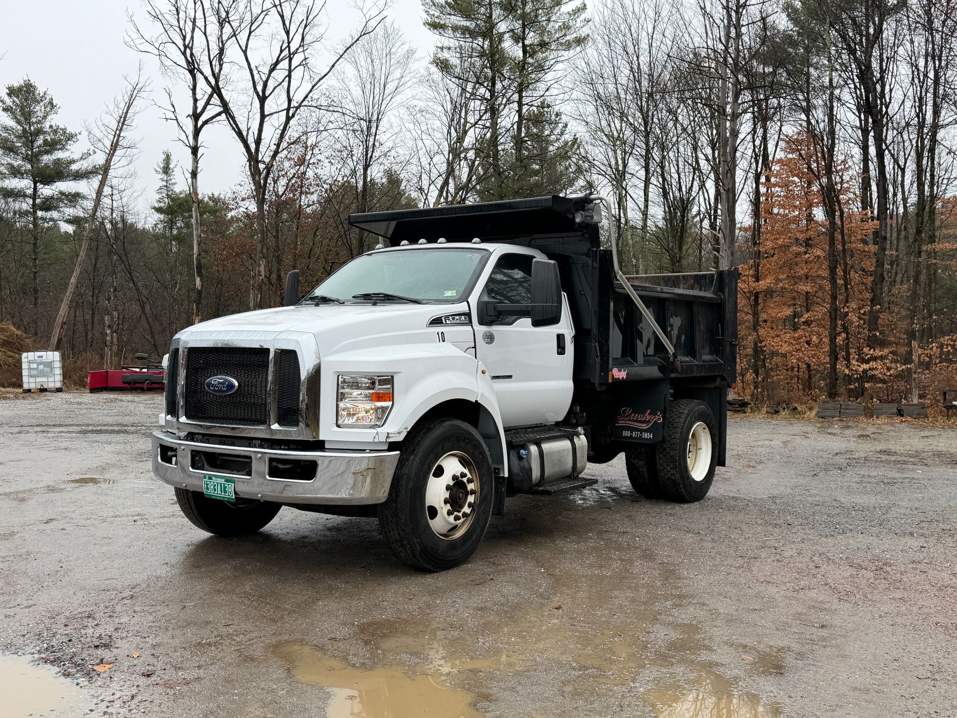 White dump truck parked on a wet, gravel area in front of trees.