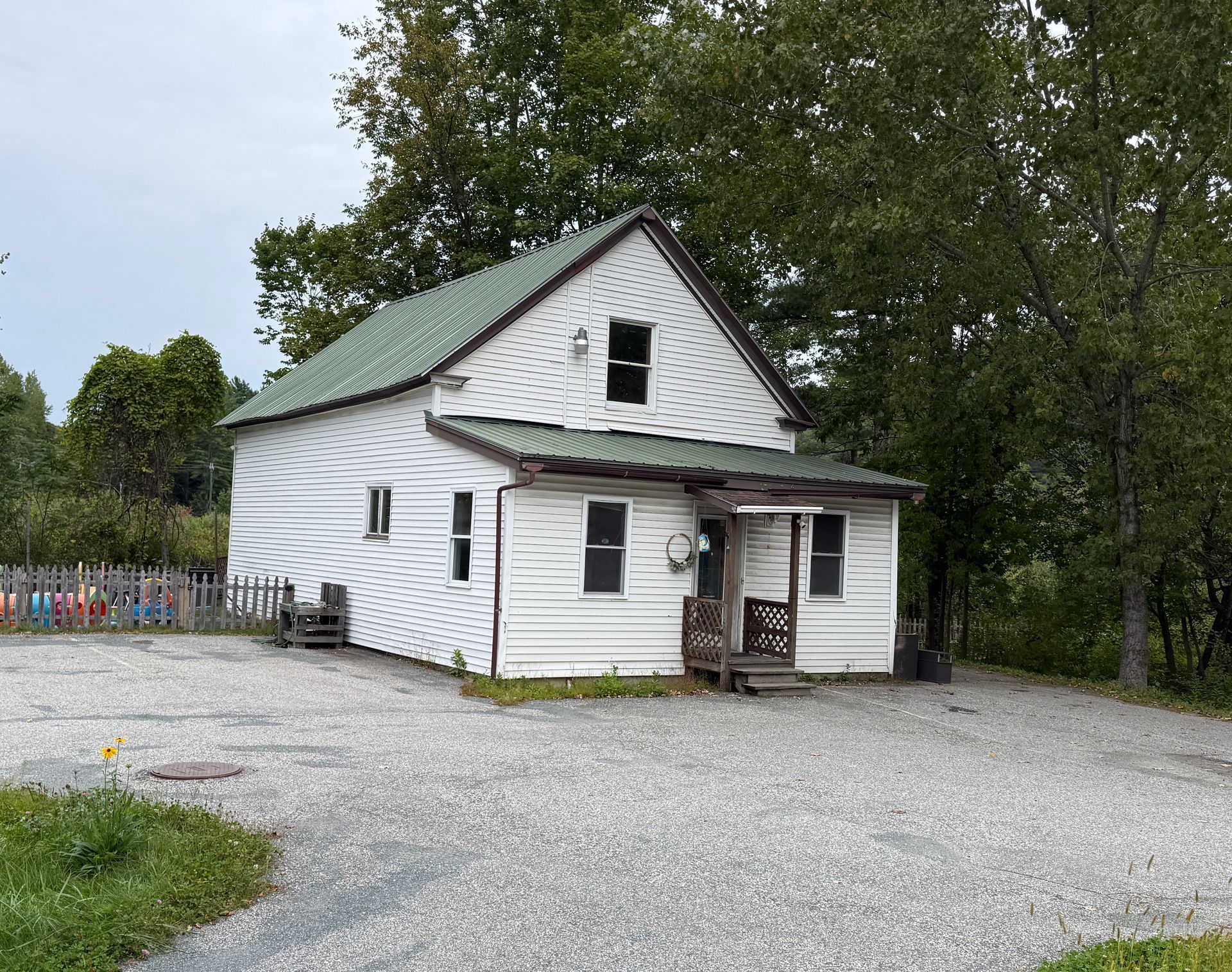 Small white house with green roof and porch, surrounded by gravel and trees
