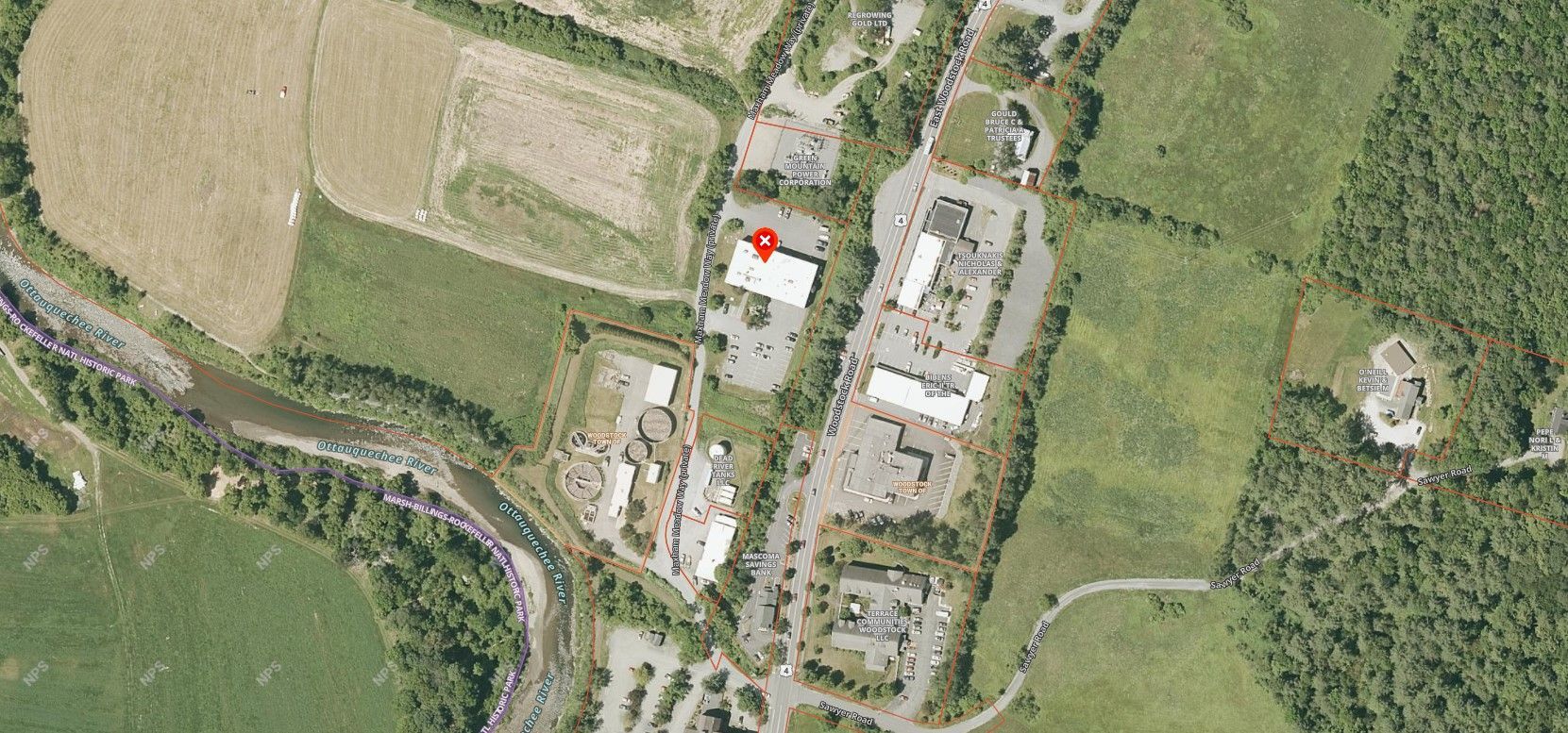 Aerial view of a commercial area with buildings, parking lots, and green spaces, including a river and forested areas.