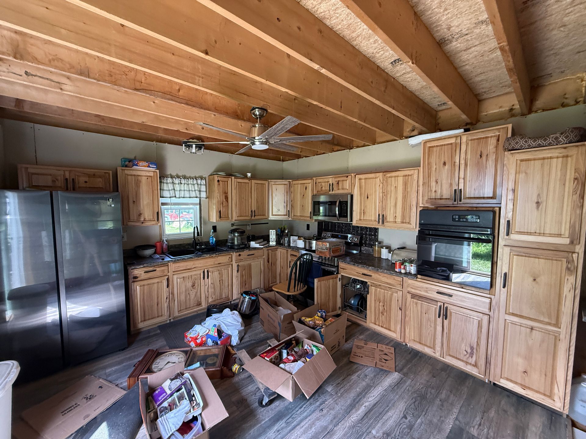 Wood-paneled kitchen with exposed beams, wooden cabinets, black appliances, and boxes on the floor