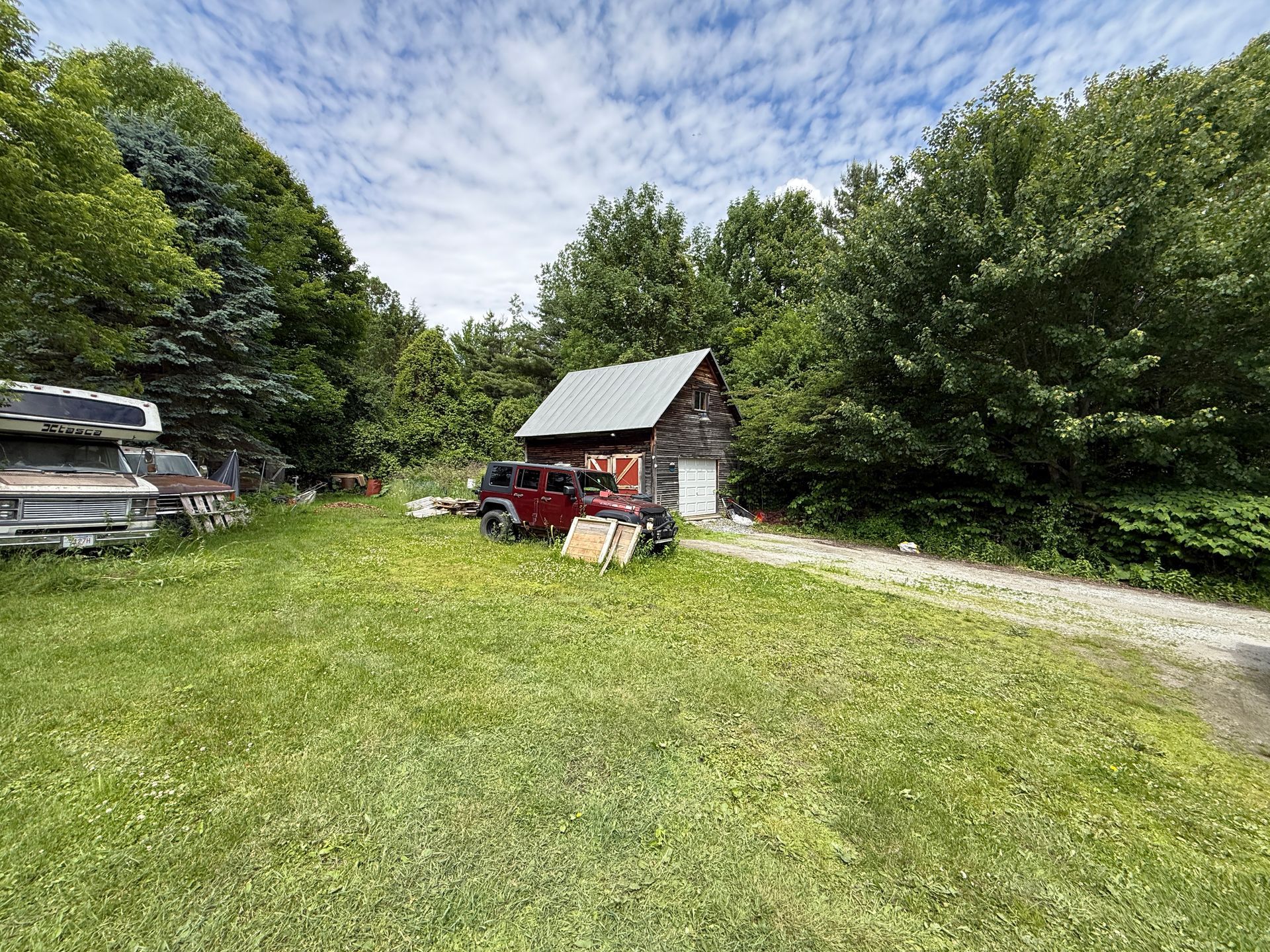 Small red shed beside a grassy yard and gravel driveway, surrounded by trees under a cloudy sky.