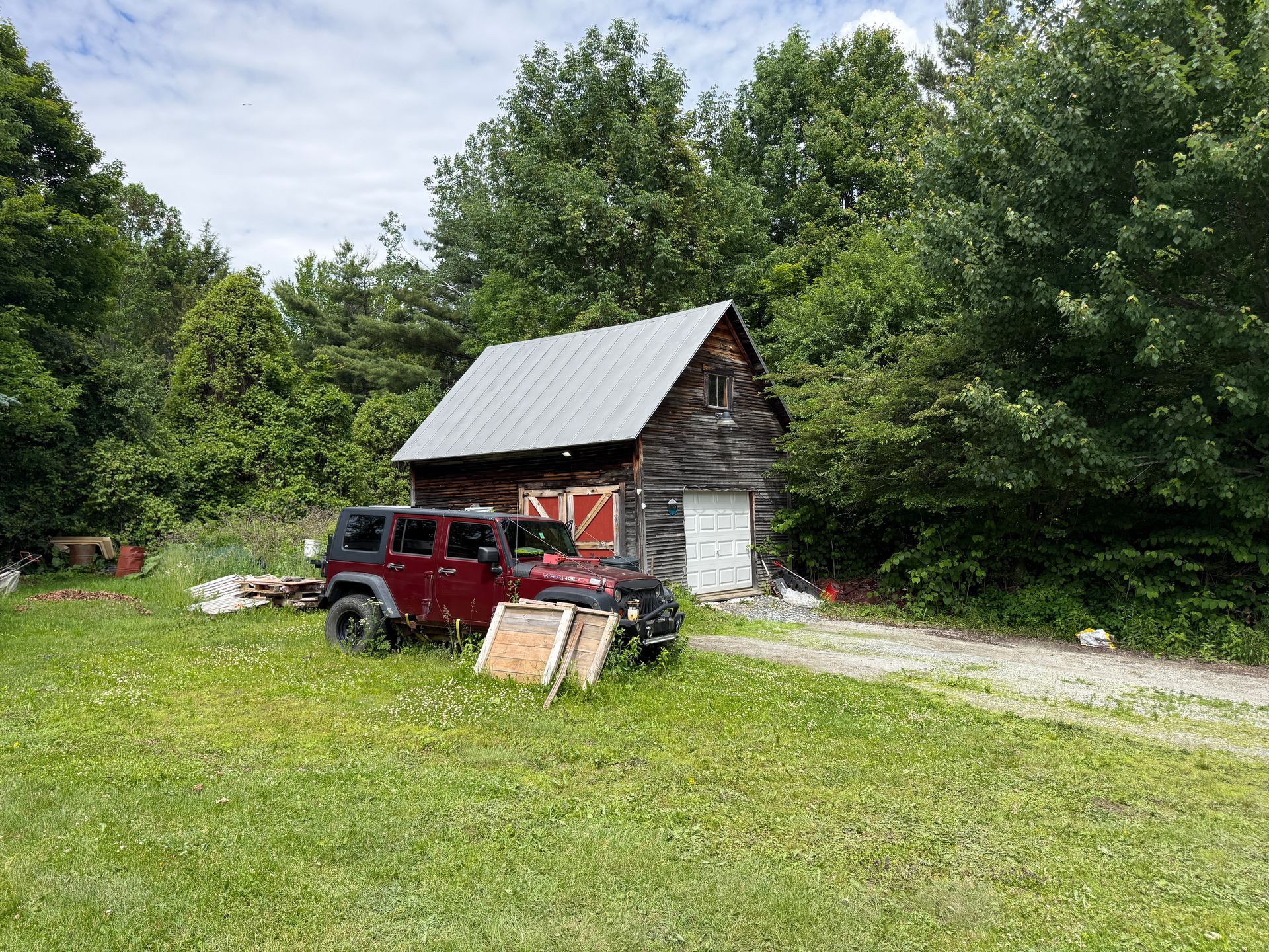 Red Jeep parked beside a small rustic barn in a grassy yard, surrounded by dense green trees.
