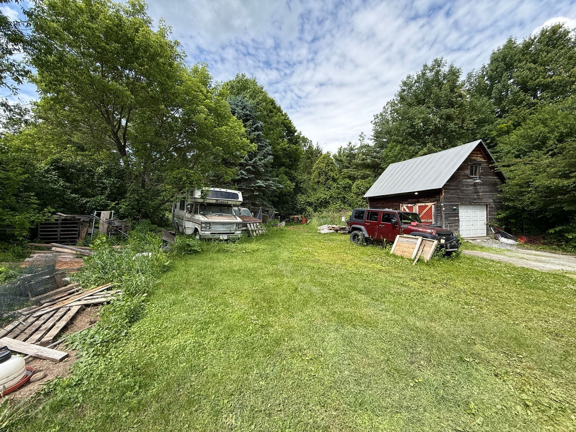 Grassy yard with trees and a small shed or barn with a red tractor under a partly cloudy sky