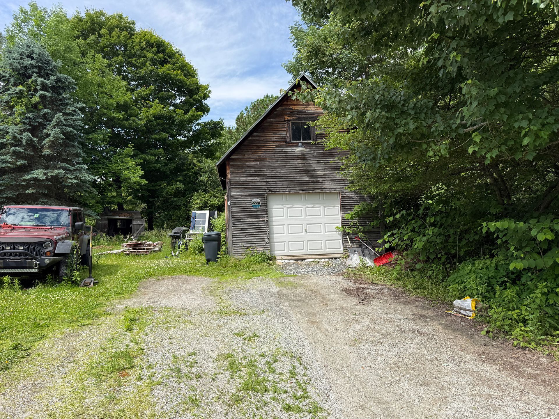 Gravel driveway leading to a rustic wooden garage surrounded by trees and greenery