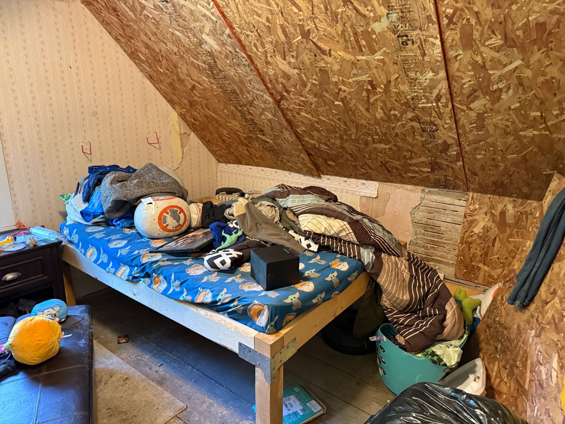 attic bedroom with a blue bed, clothes, and slanted plywood walls.