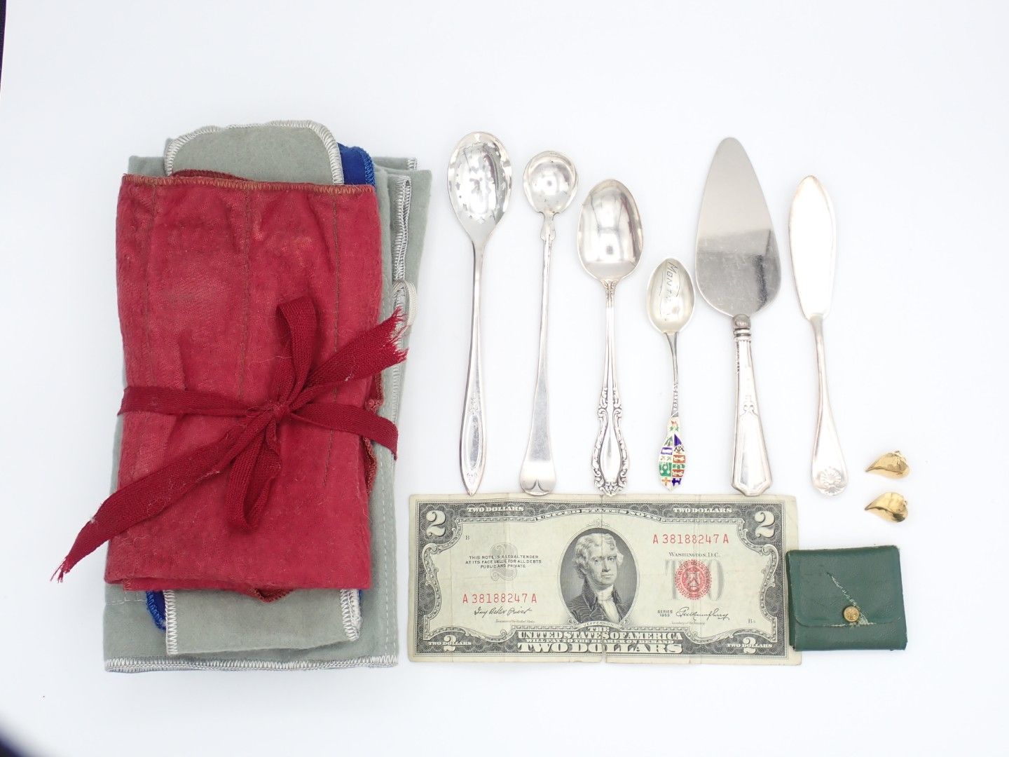 Small wrapped gift, silver cutlery, a two-dollar bill, a green envelope, and tiny gold items on a white background