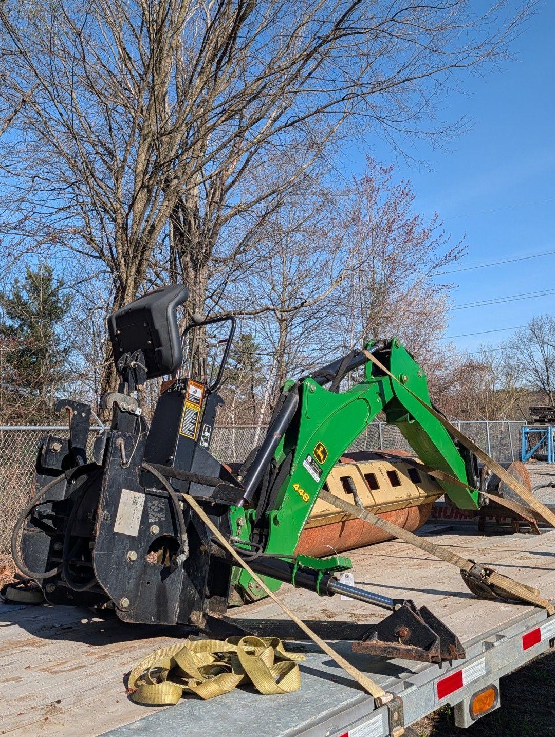 Green excavator and black skid steer on a flatbed trailer at a construction site under bare trees