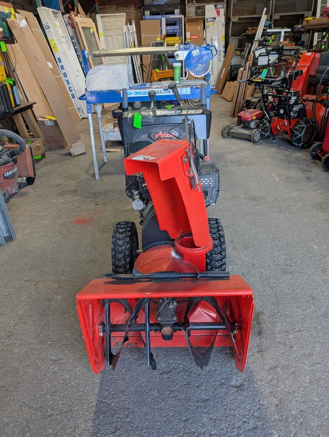 Red snow blower in a workshop, front-facing with black wheels and a blue engine on top.