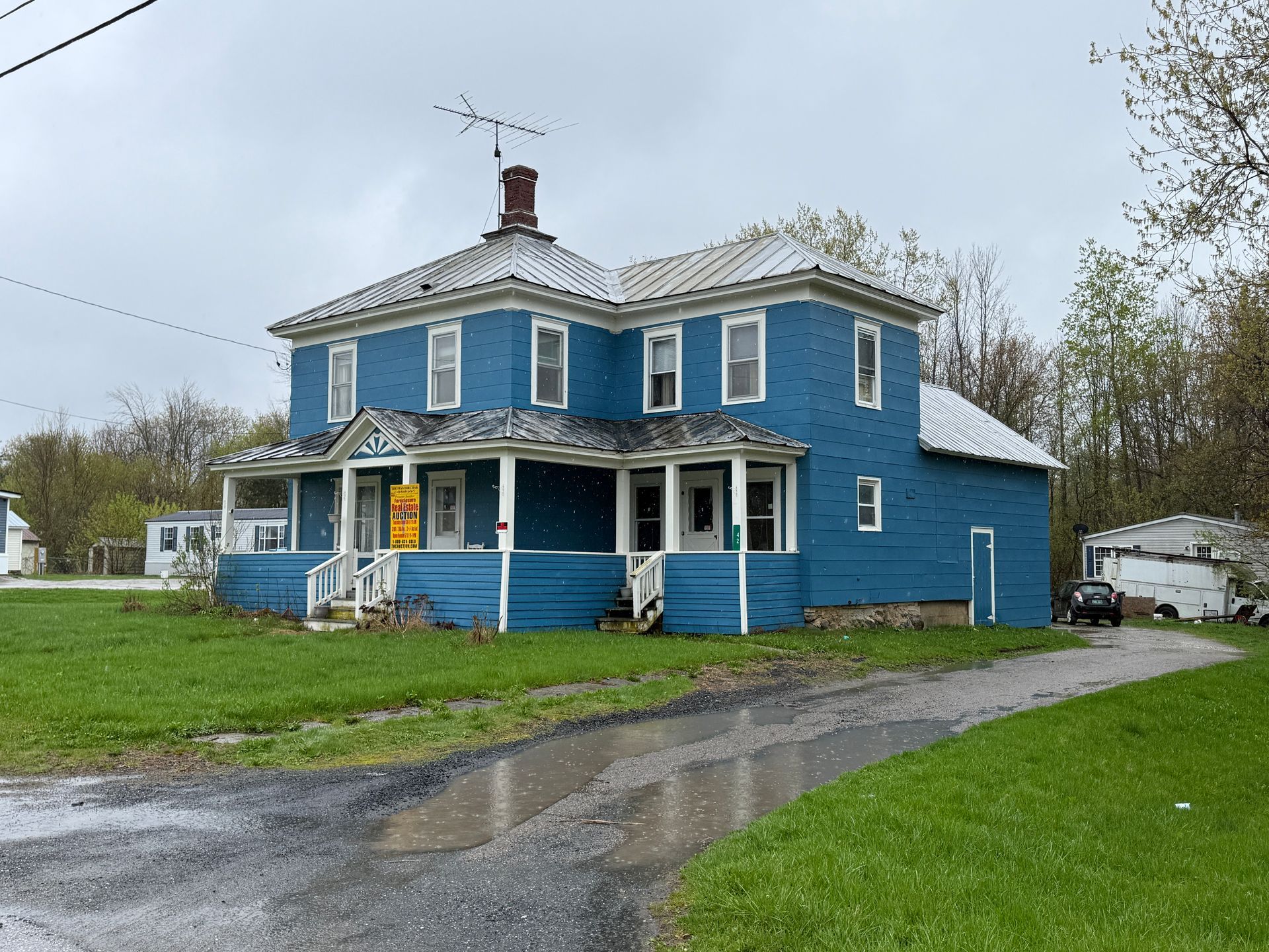 Blue two-story house with white trim and a porch, beside a wet driveway on a cloudy day