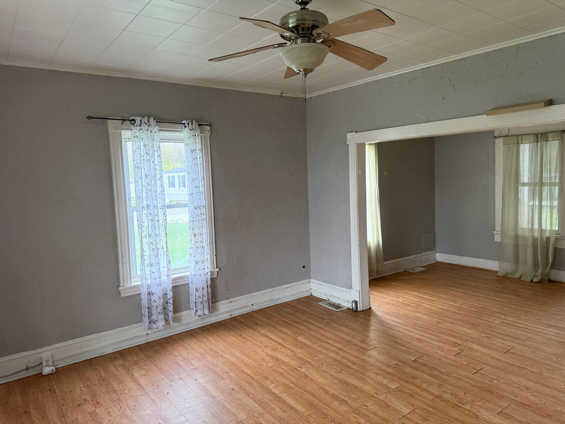 Empty room with gray walls, wood floors, ceiling fan, and a wide doorway to an adjoining room.