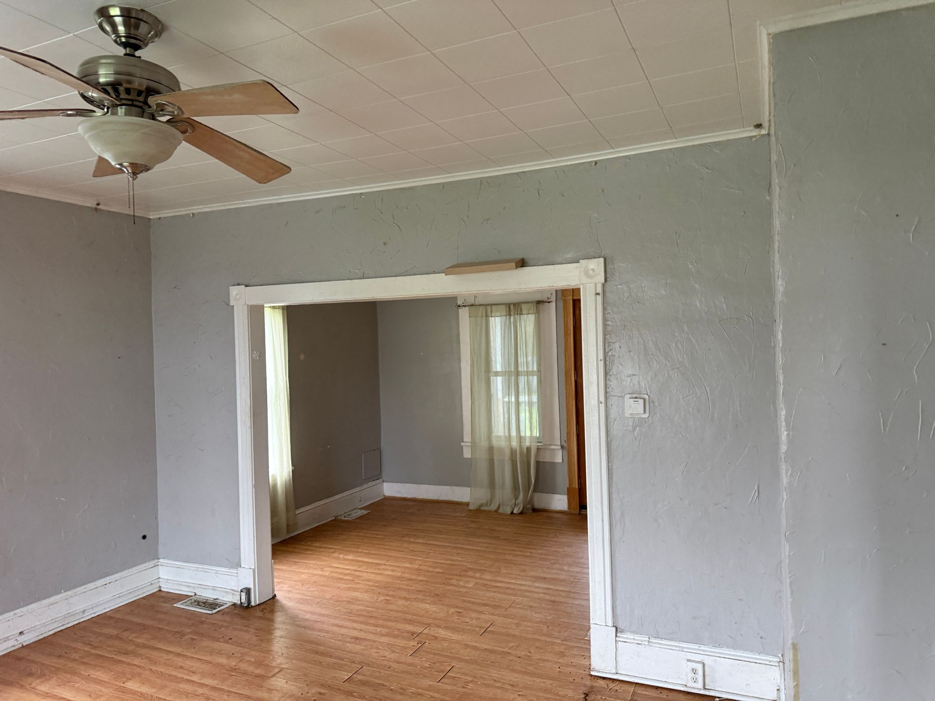 Empty gray room with wood floor, ceiling fan, and open doorway to a second room with a window.