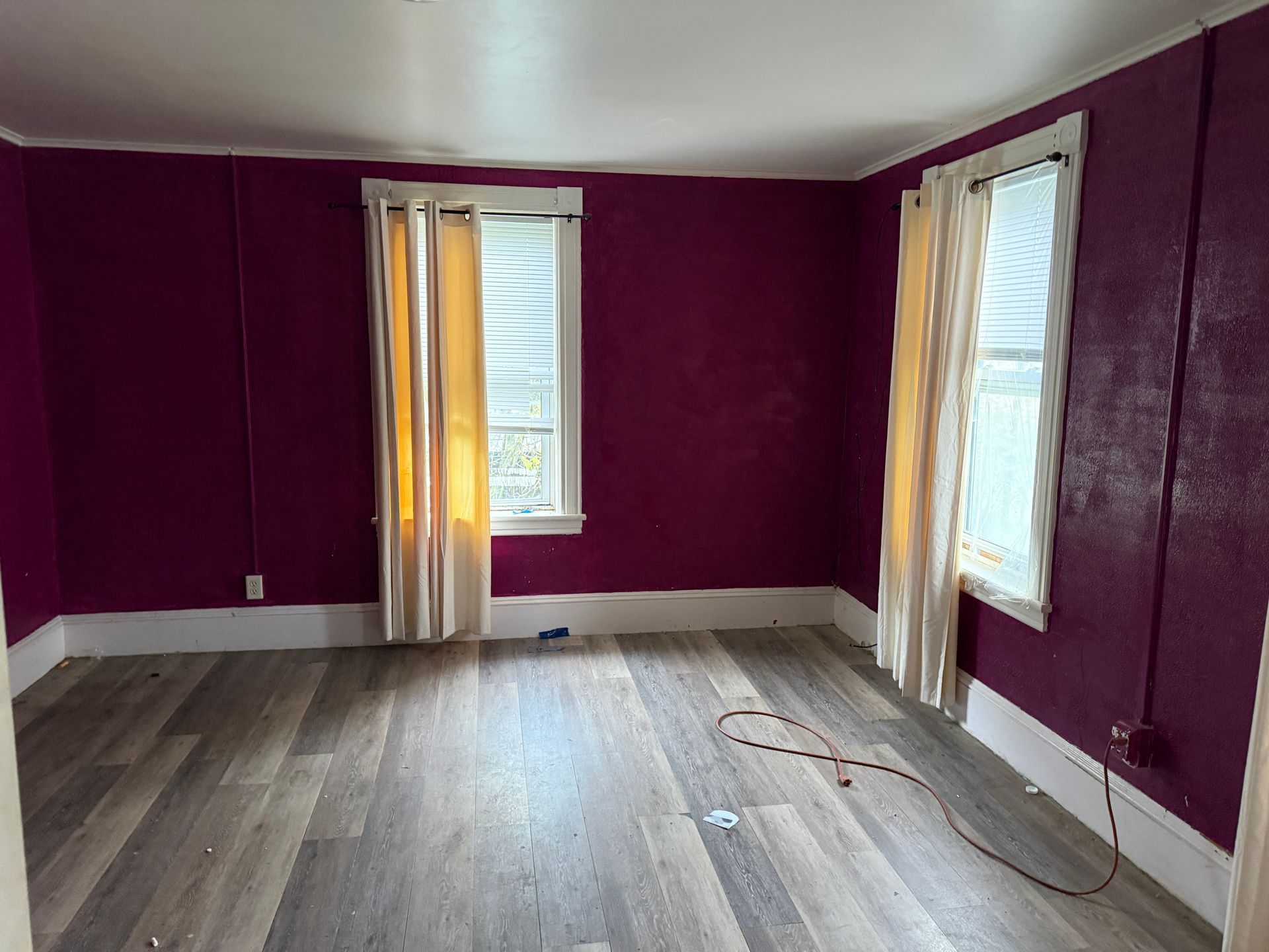 Empty room with purple walls, gray wood floors, and two tall windows with white curtains.
