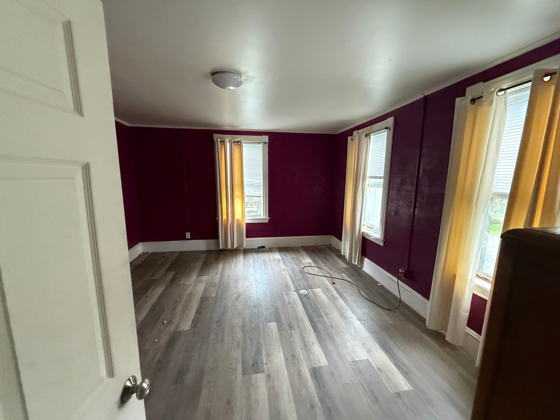 Empty room with burgundy walls, gray wood floors, and tall windows with yellow curtains.