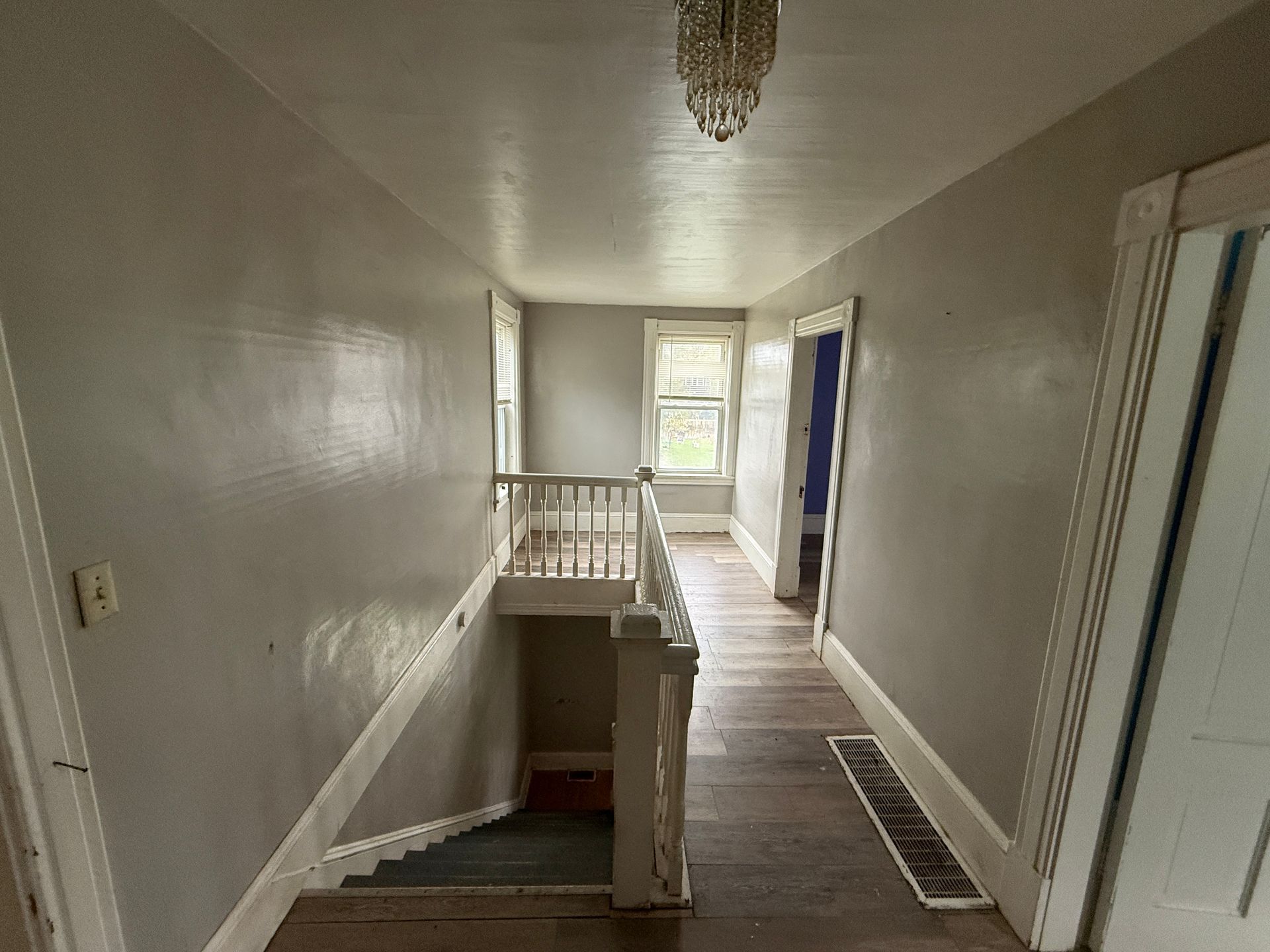Hallway with white walls, staircase railing, chandelier, and doors leading to a bright room.