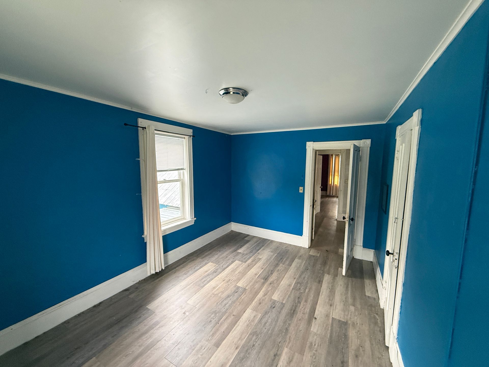 Empty bright blue room with gray wood flooring, white trim, and a doorway to a hallway