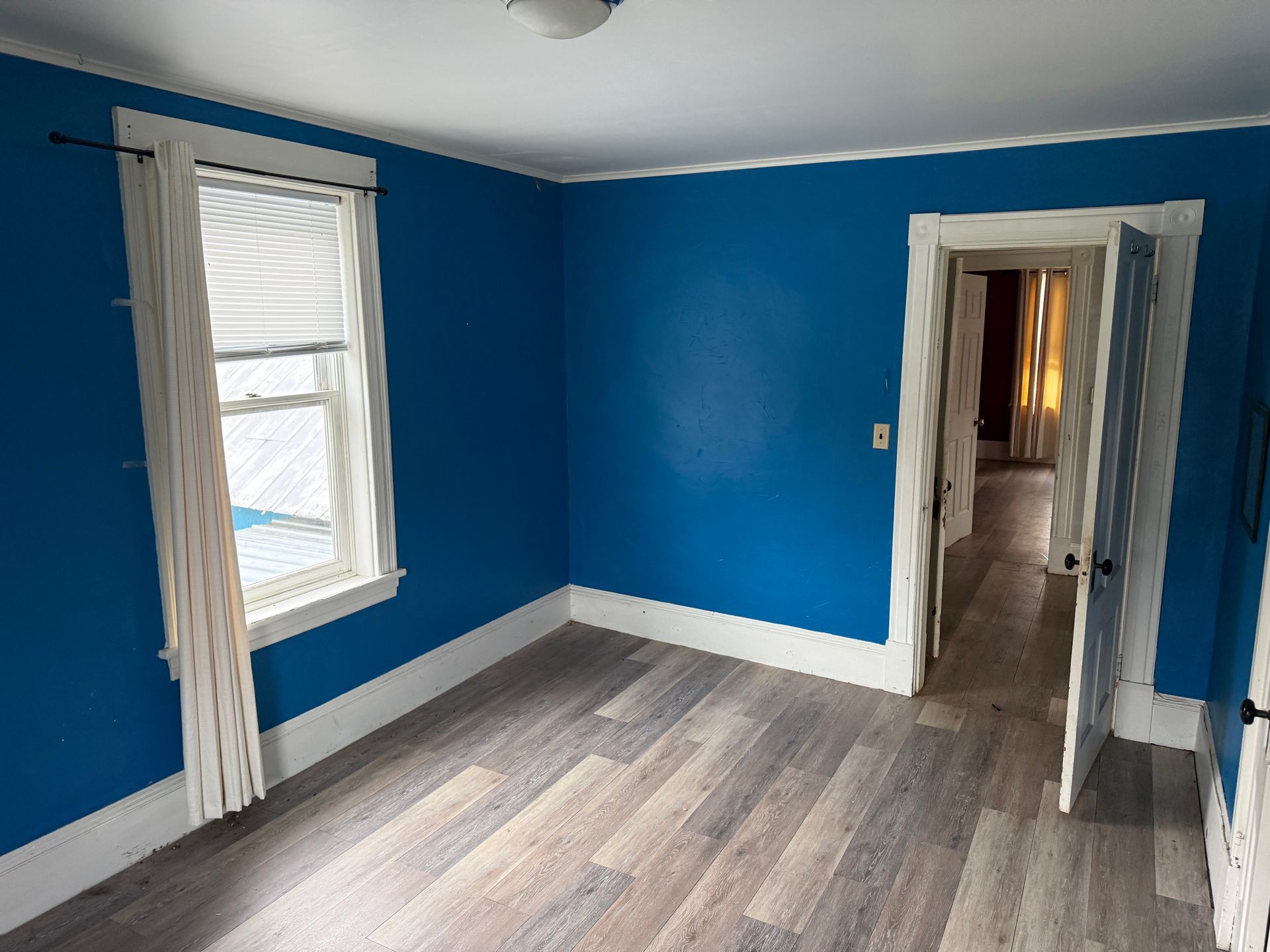 Empty room with bright blue walls, gray wood flooring, a window, and an open doorway to a hallway