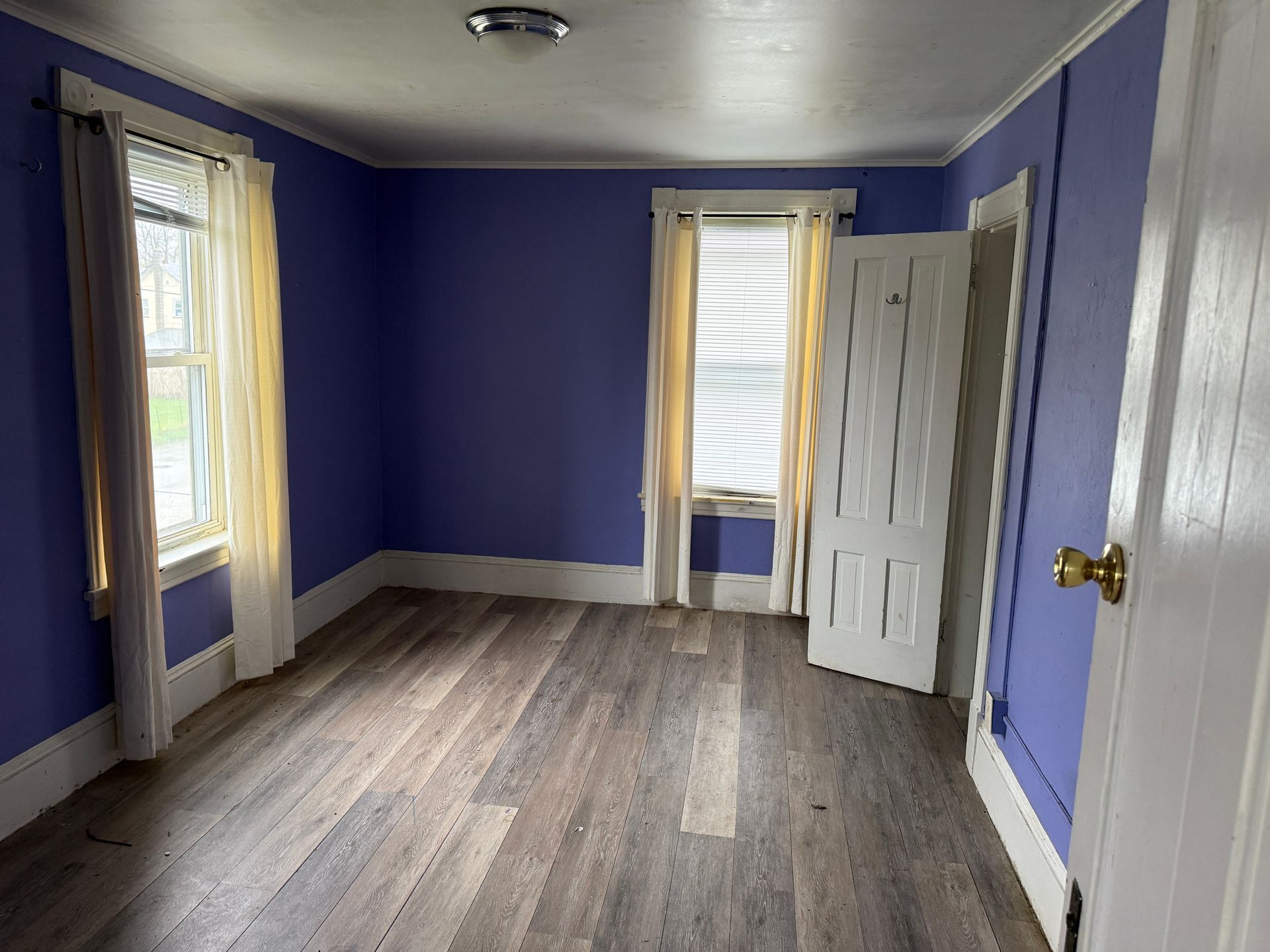 Empty room with purple walls, wood floor, and windows with curtains and blinds