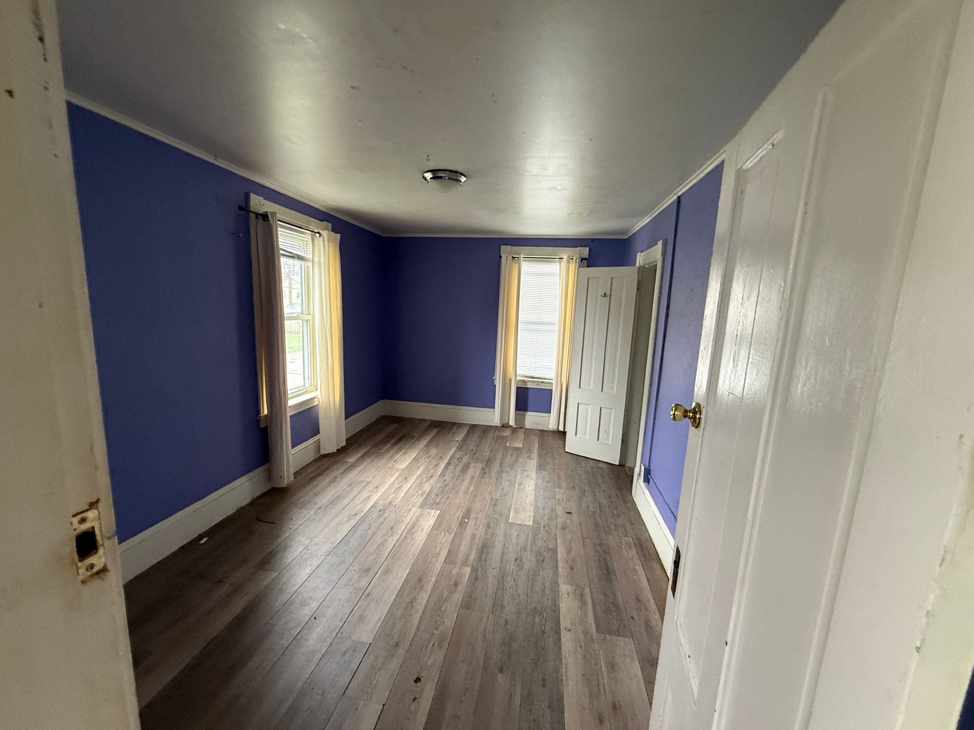 Empty room with blue walls, wood floor, and white closet doors, lit by two windows.