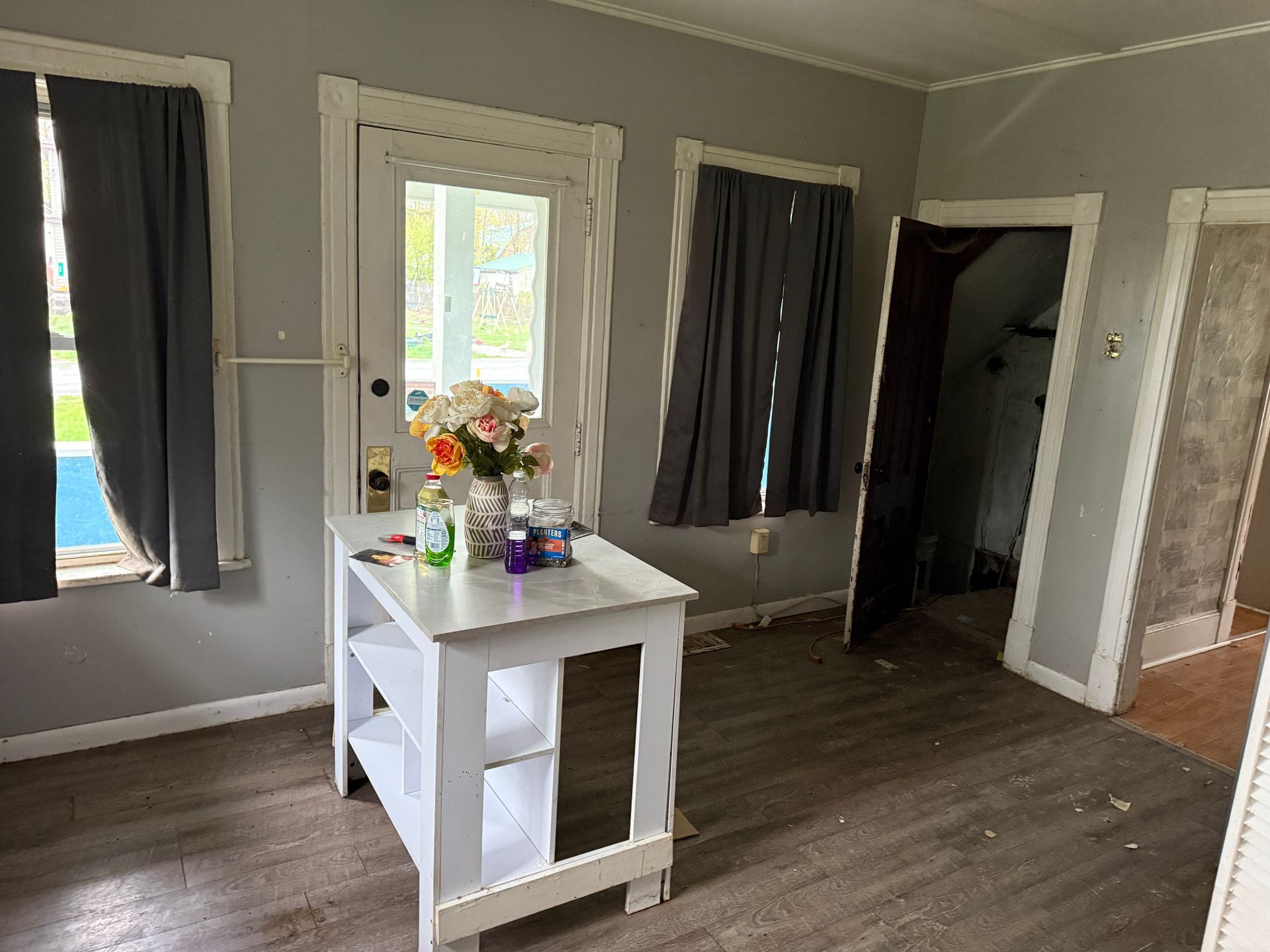 Small room with white walls, dark wood floors, curtained windows, and a white table holding flowers and bottles