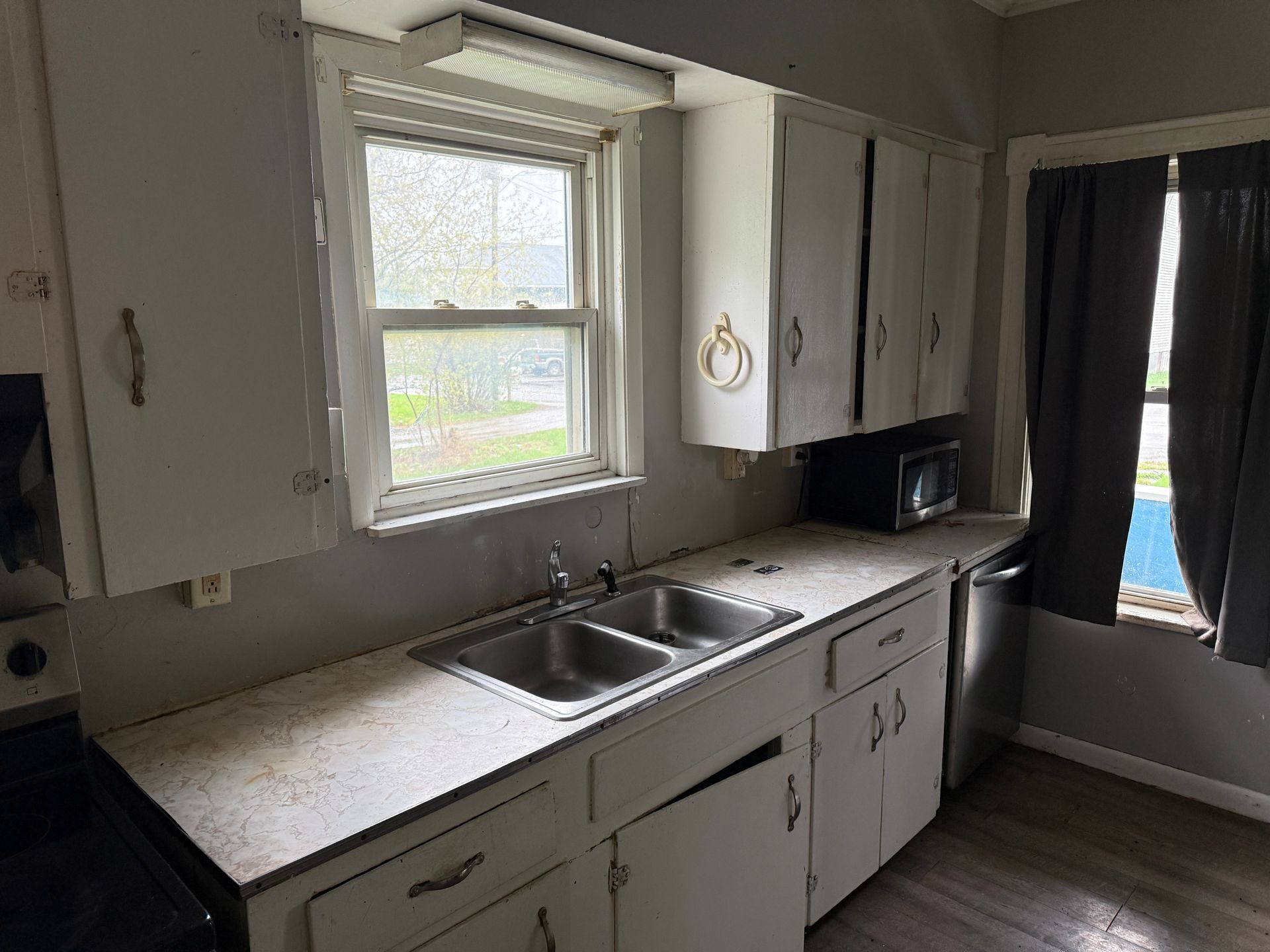 Small kitchen with white cabinets, double sink, window, and dark curtains by a blue door.