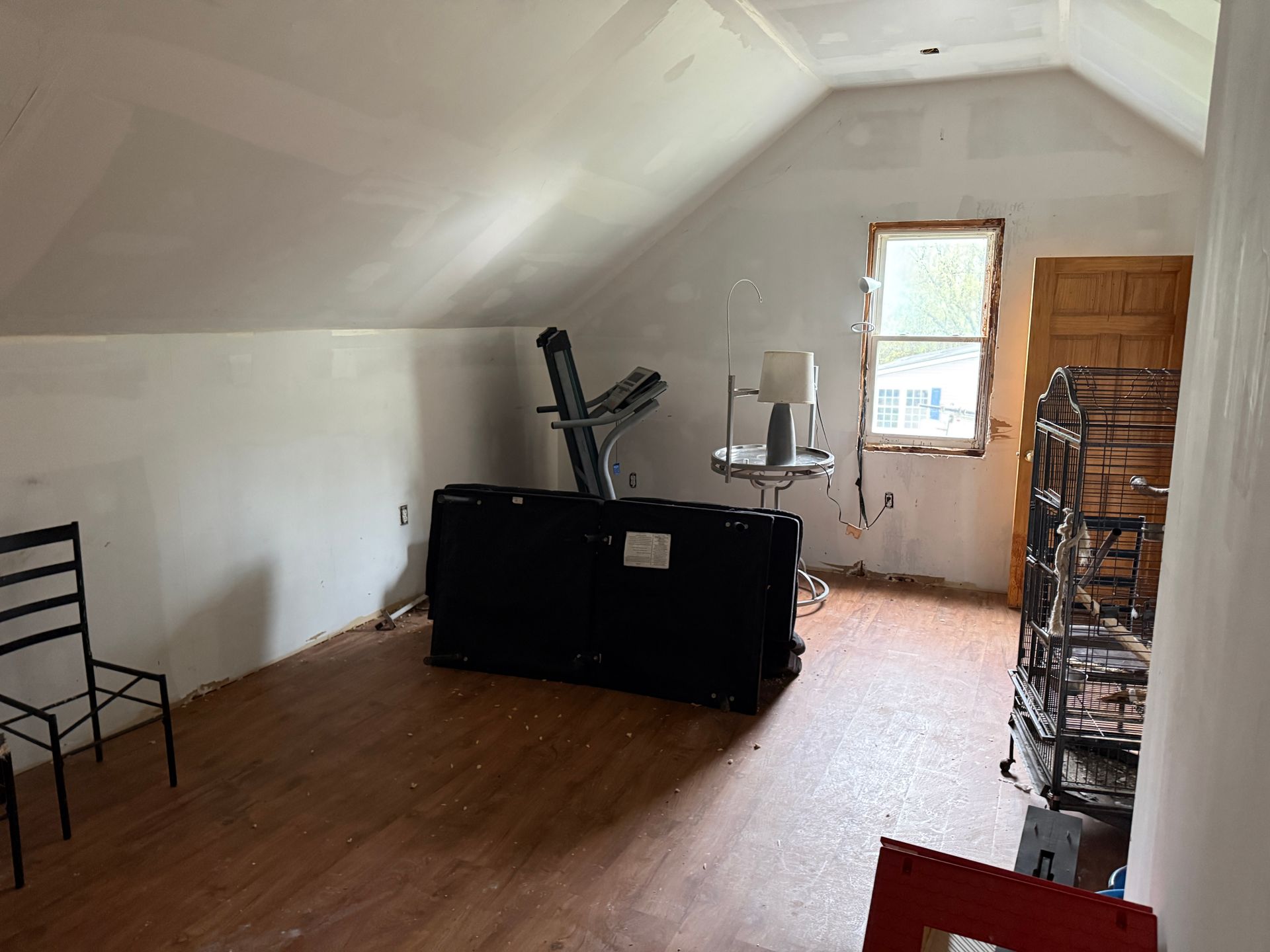 Empty attic room with wood floor, black furniture, and a small window at the far end.