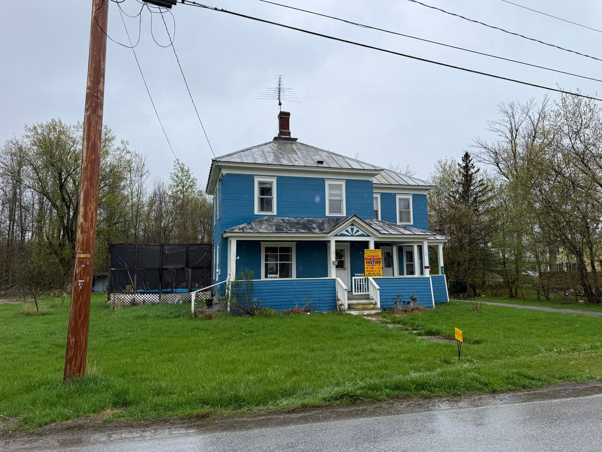 Blue two-story house with a porch beside a road, with grass, trees, and a utility pole in front