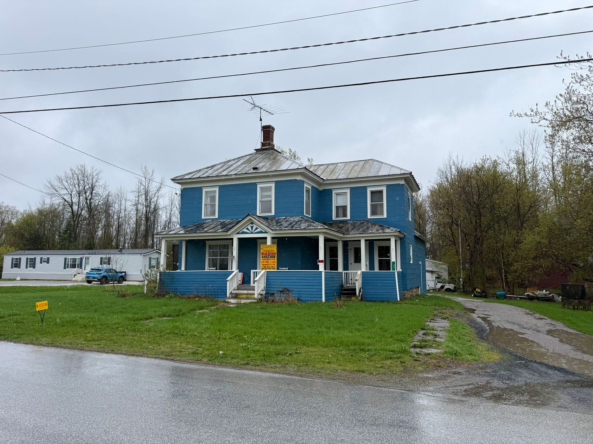 Blue two-story house with a porch on a wet roadside under cloudy skies