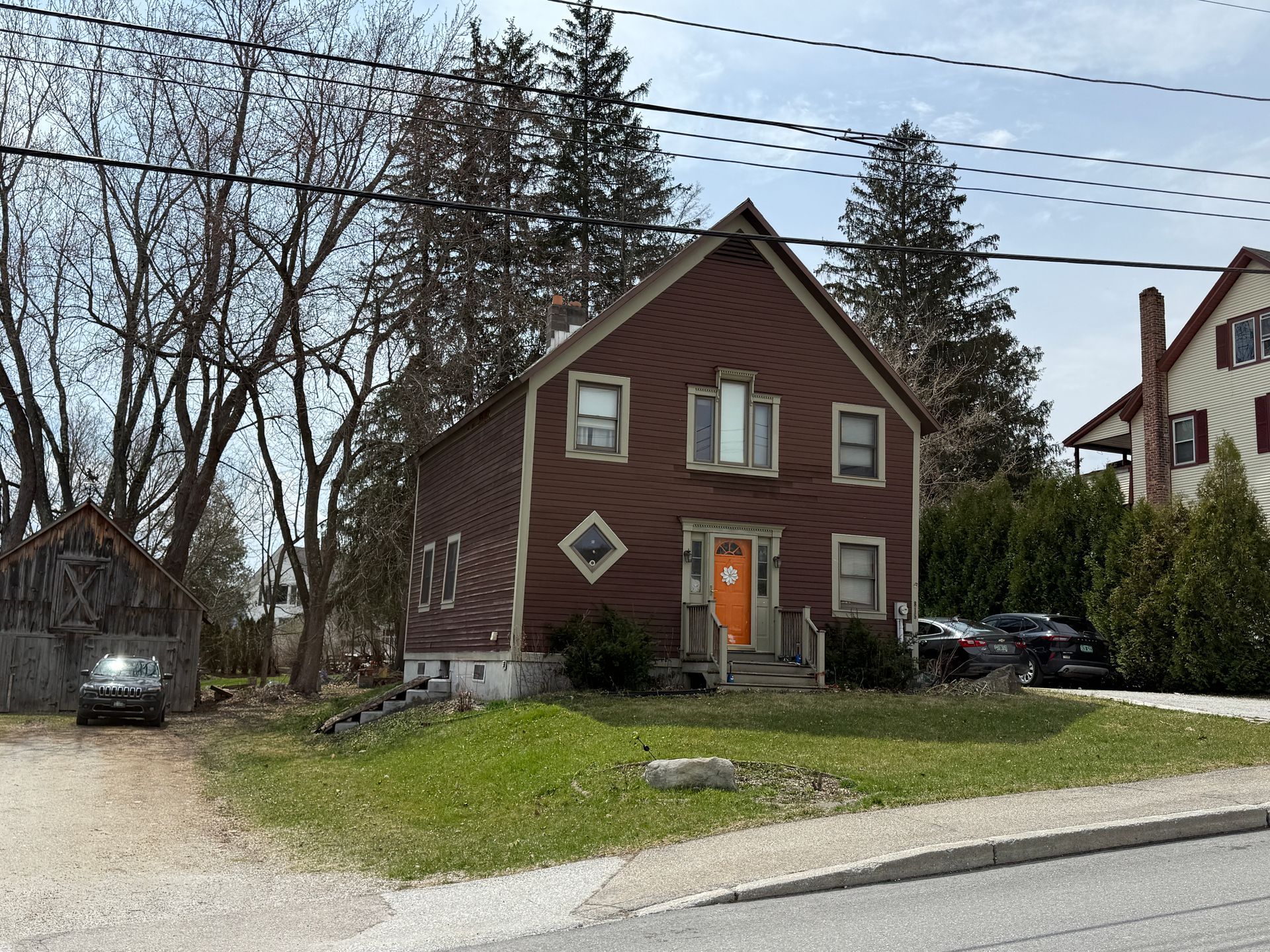 Small brown house with an orange door beside a snowy roadside, with trees and parked cars nearby.