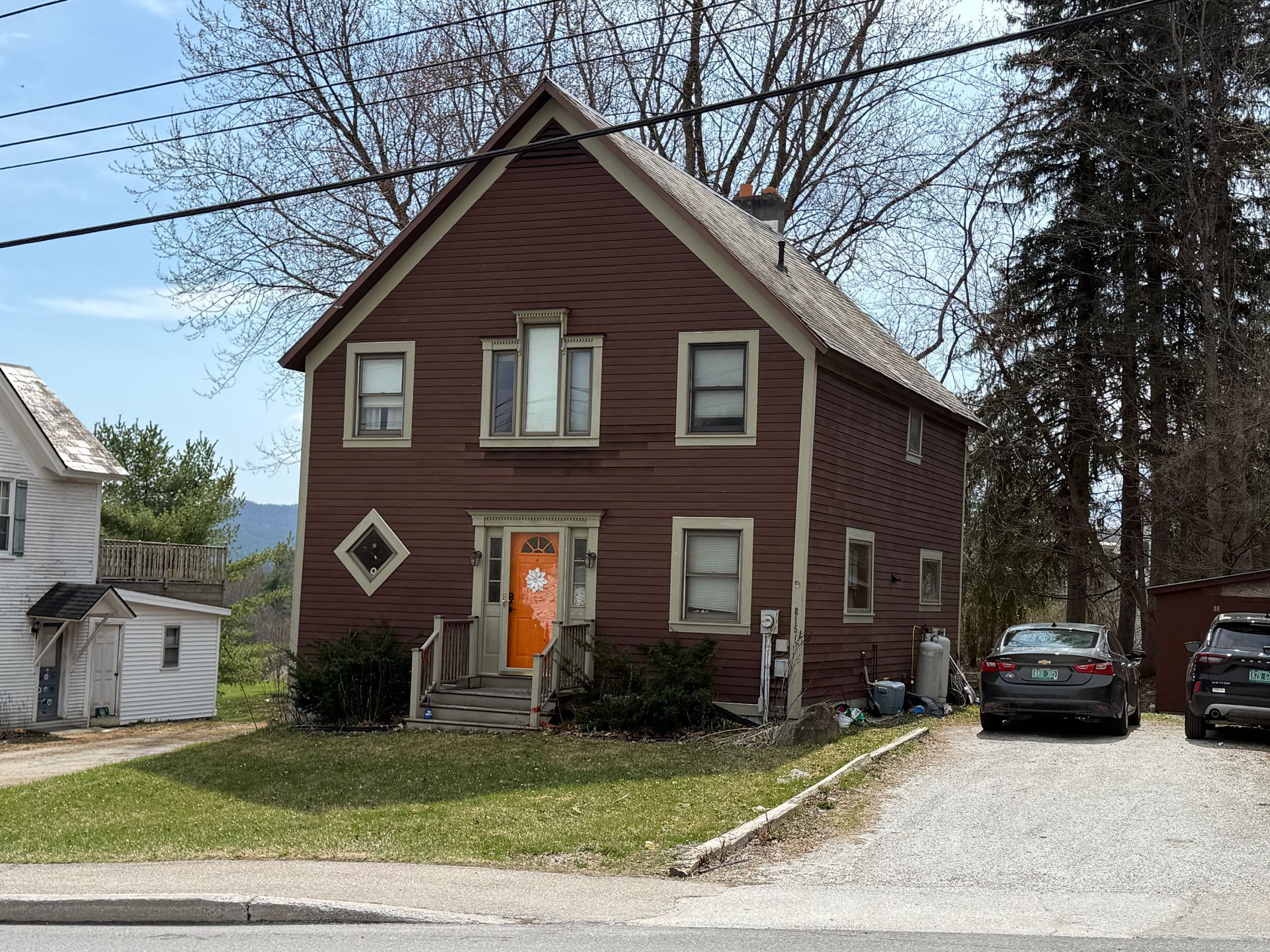 Two-story brown house with an orange front door, diamond window, and cars parked on the side drive.