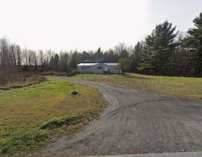 Gravel driveway curving toward a white house, with grassy fields and trees under a bright sky