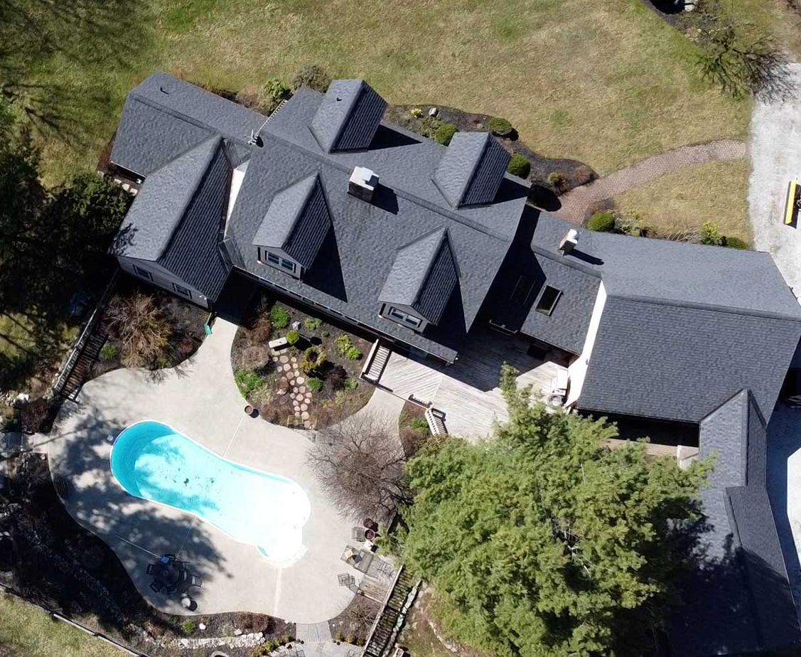 Aerial view of a house with a dark roof and a pool. Includes a driveway and a large tree.