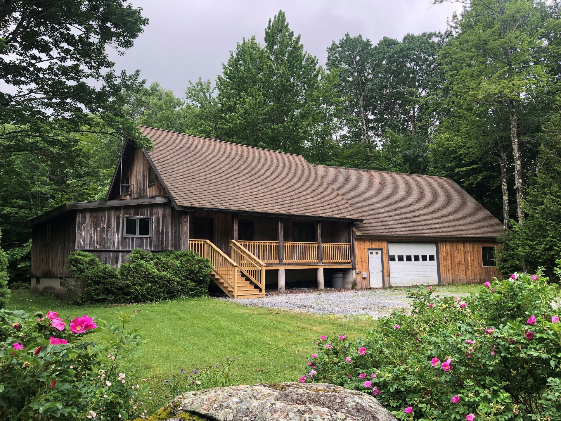 Rustic wooden cabin with porch, garage, and a lush green yard, surrounded by trees and flowers.