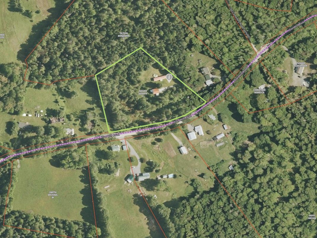 Aerial view of a rural hillside with scattered houses, fields, and a winding road through dense green forest.
