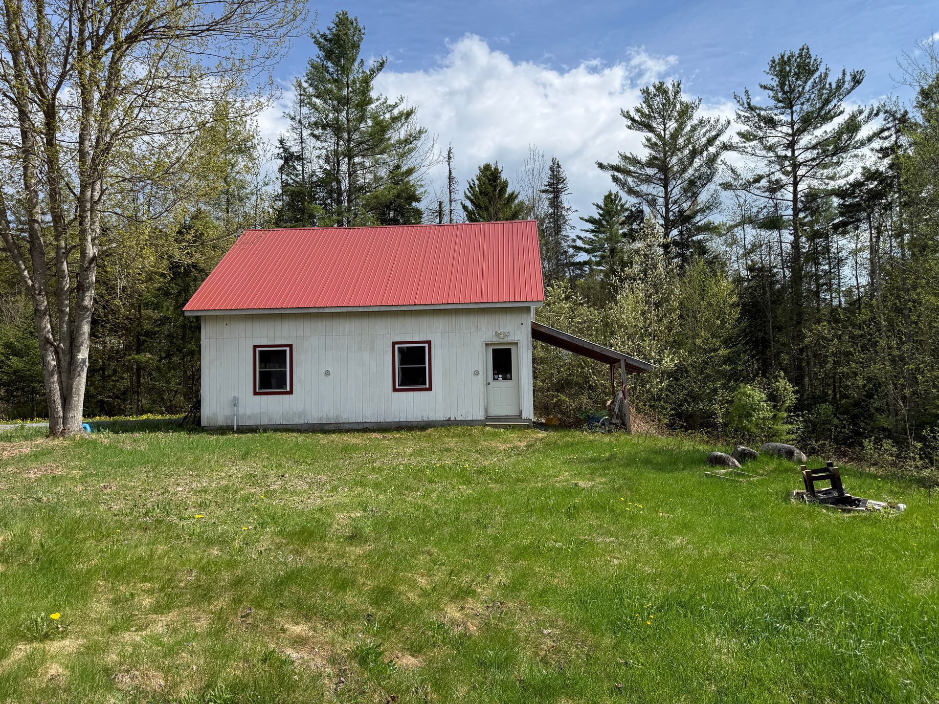 Small white shed with red roof on a grassy yard, surrounded by trees under a partly cloudy sky.