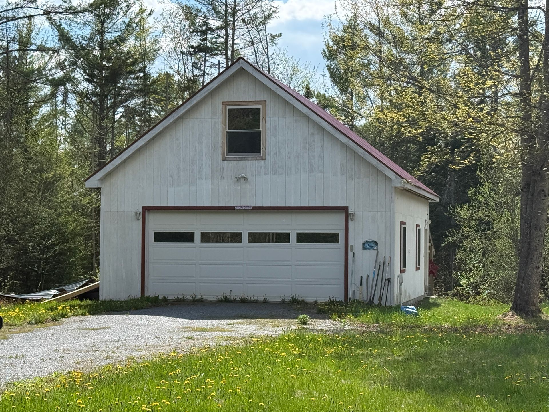 White garage with a gabled roof and single window, set beside a gravel drive and grassy yard among trees.
