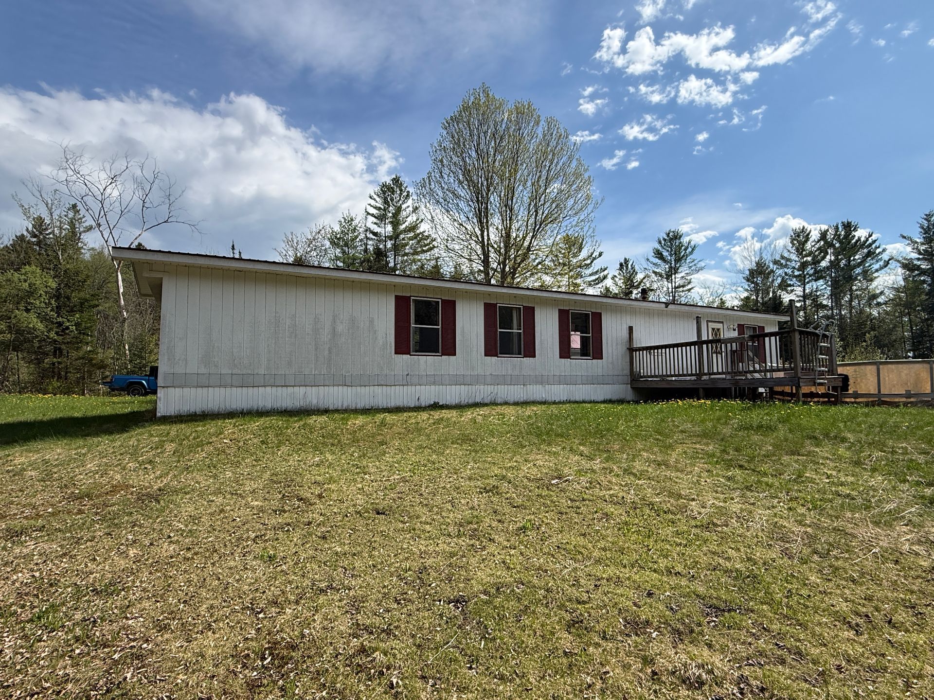 Single-story white house with red shutters beside a wooden deck in a grassy yard under a blue sky