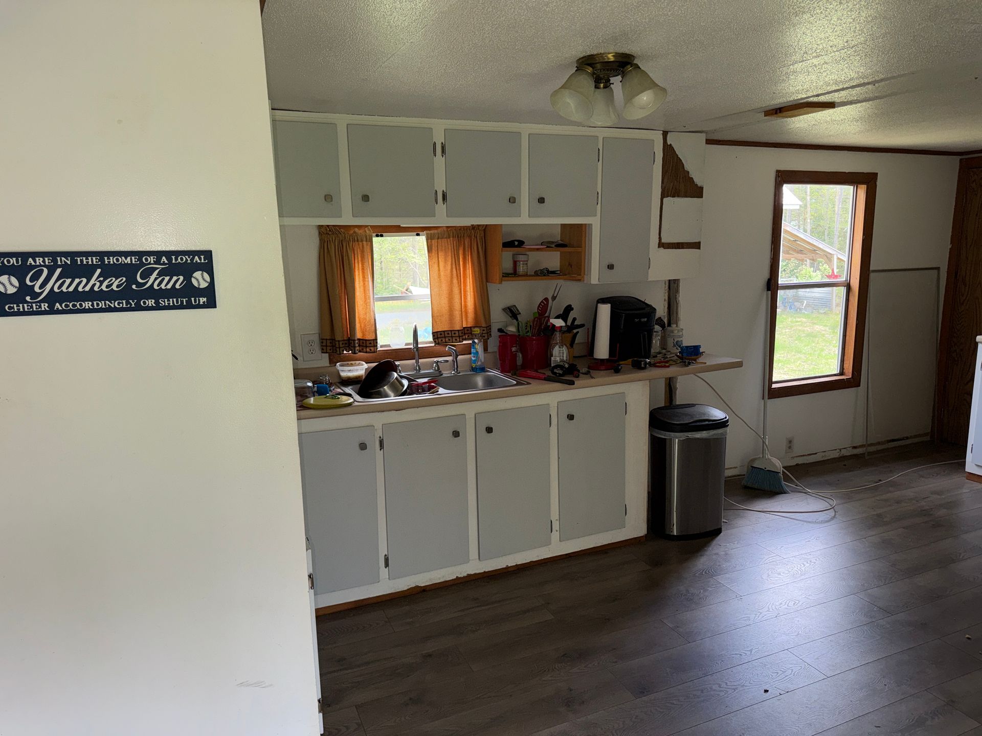 Small kitchen with white cabinets, wood floor, sink, trash can, and a window overlooking a yard.