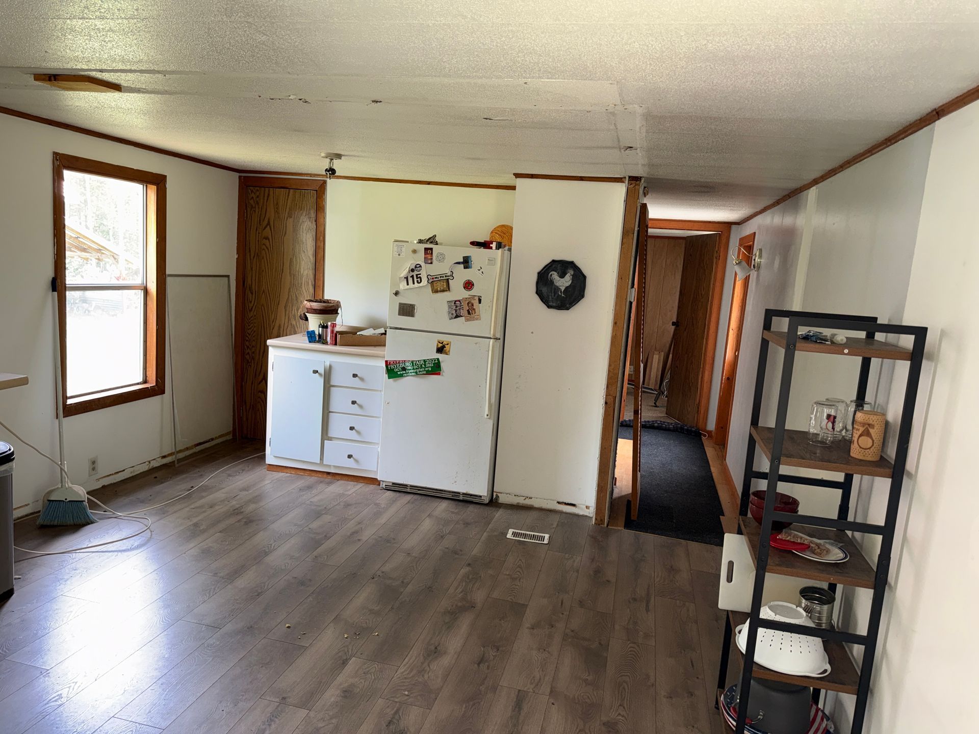 Empty living room with wood floors, a white cabinet and fridge, and a black shelving unit near a hallway.