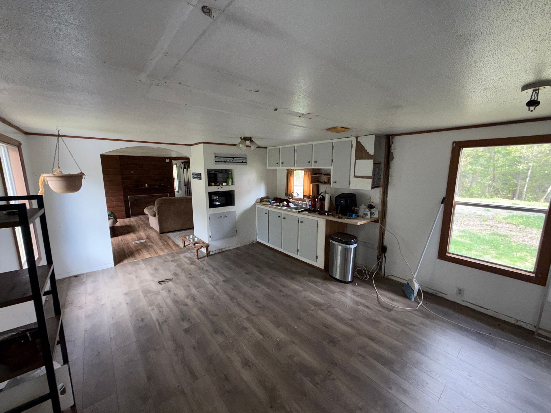 Empty rustic kitchen and living area with white cabinets, wood floors, and large window looking outside