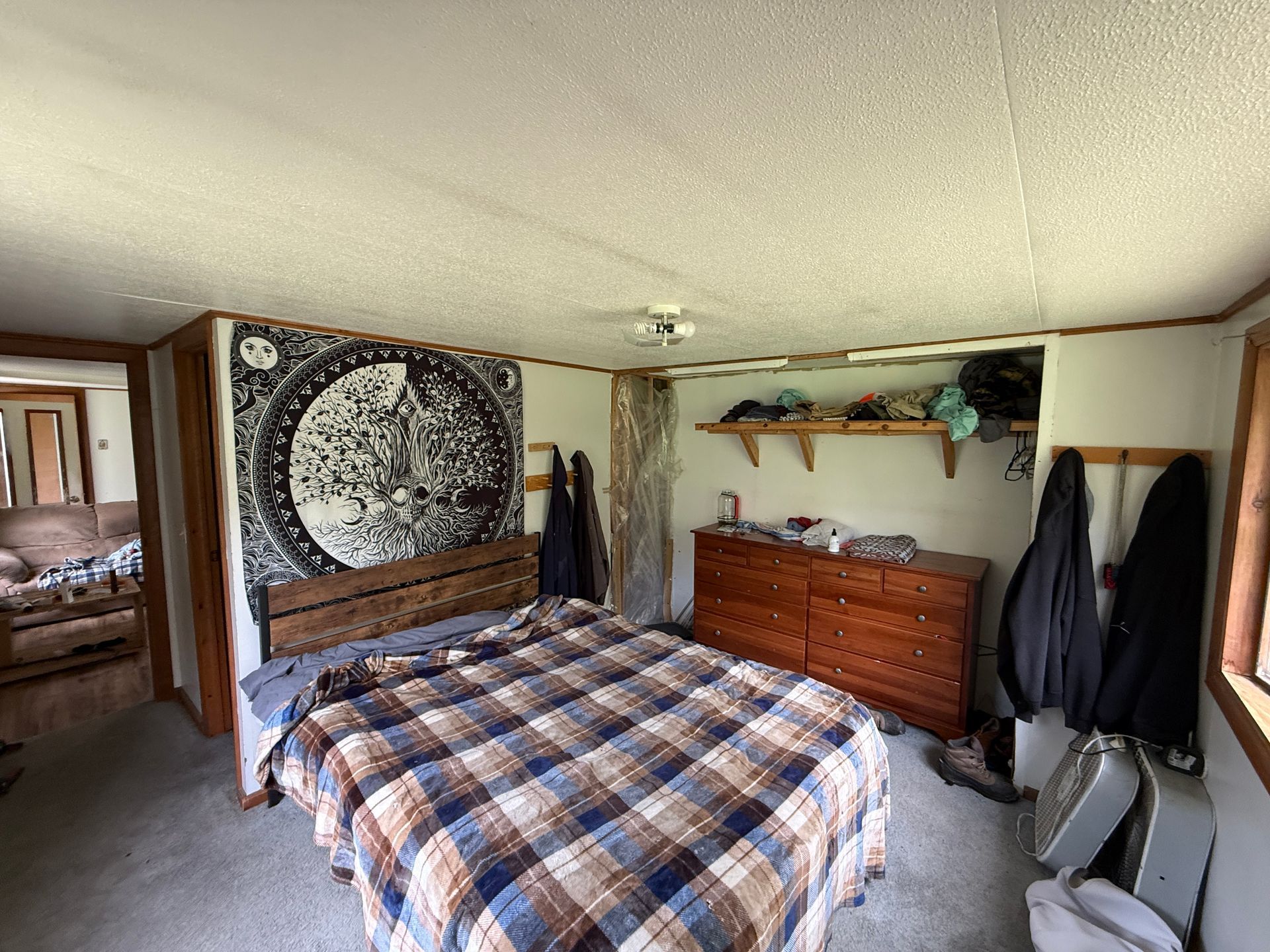 Bedroom with plaid bedspread, tapestry wall art, dresser, coat rack, and doorway to another room